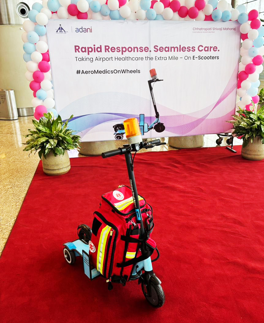E-scooter with a red medical bag on a red carpet, promoting Adani's "AeroMedicsOnWheels" airport healthcare service.