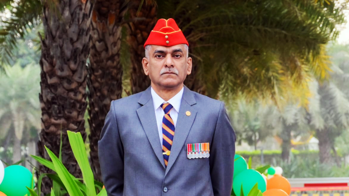 Portrait of a man in a grey suit, red cap, and striped tie, adorned with medals, standing outdoors.