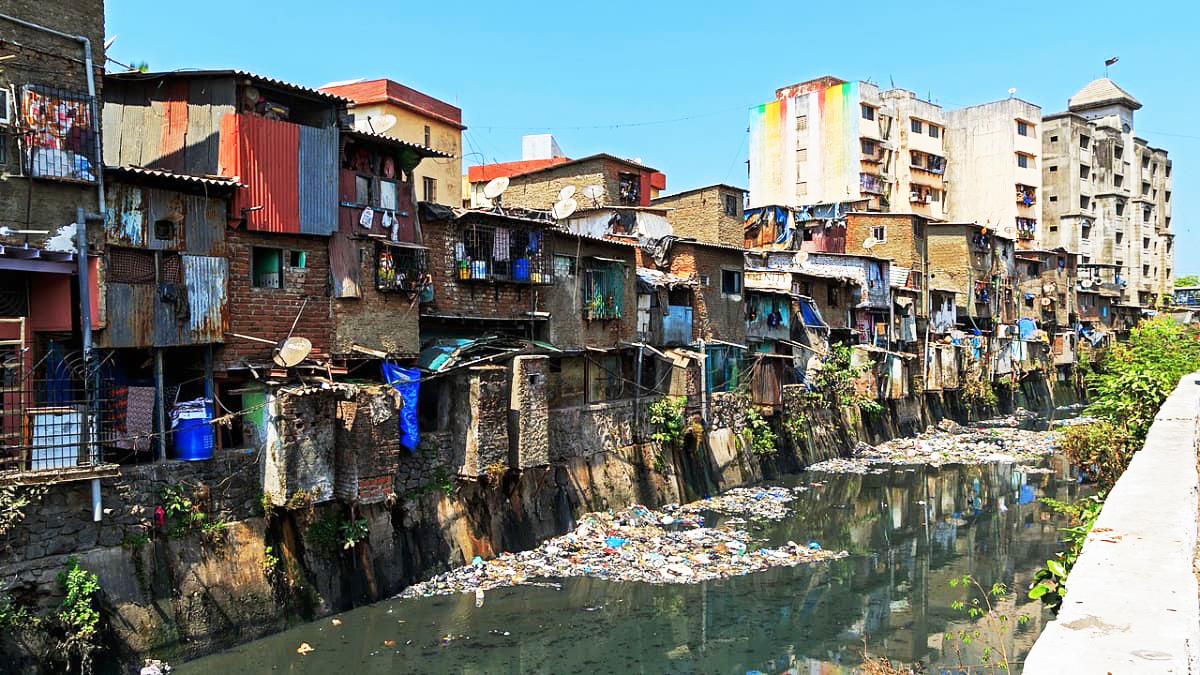 Slum dwellings with corrugated metal and brick walls lining a heavily polluted canal filled with trash under a clear sky.
