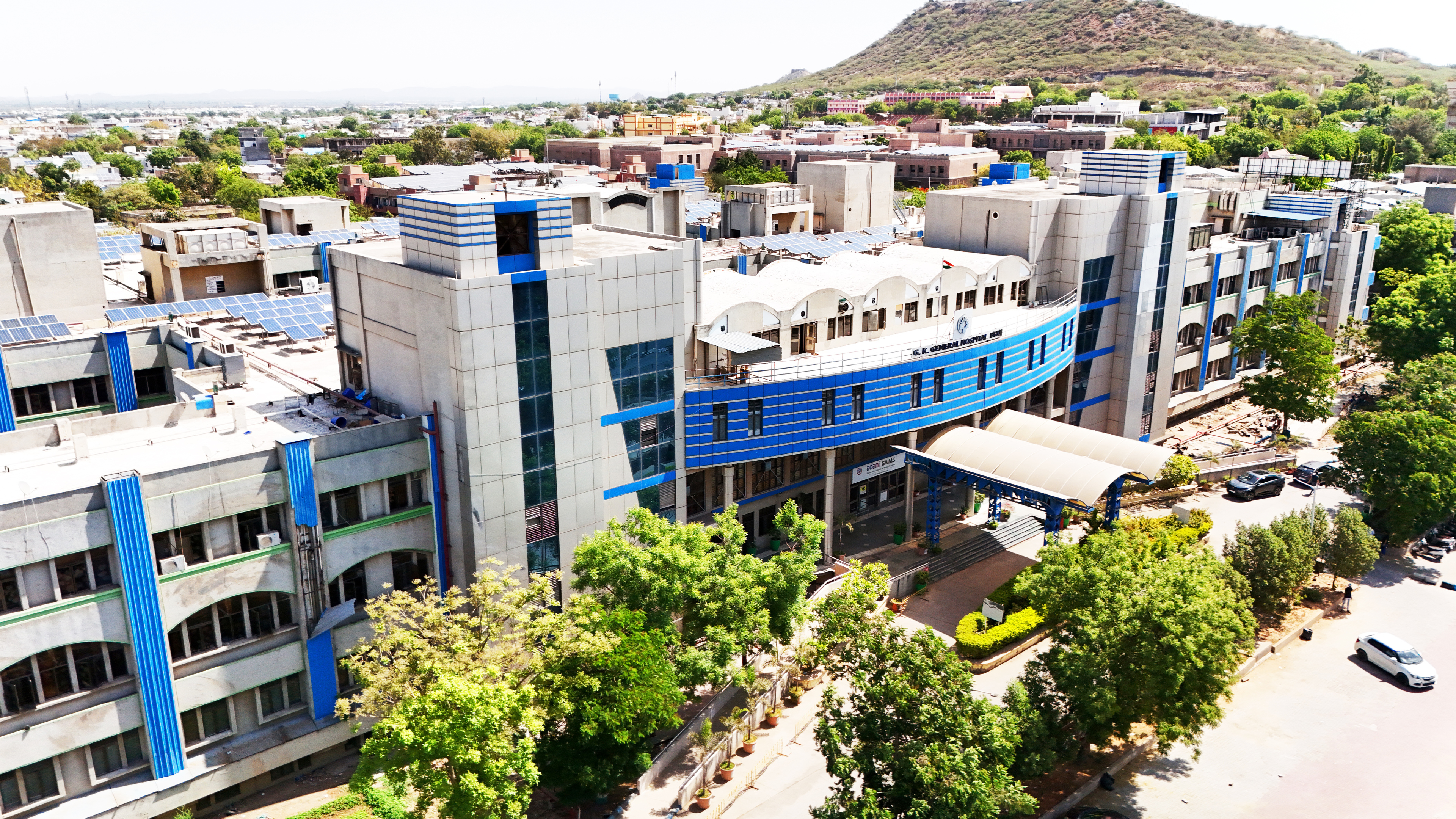 Aerial view of a modern hospital building with blue and white facades, solar panels, surrounded by a town and hills.