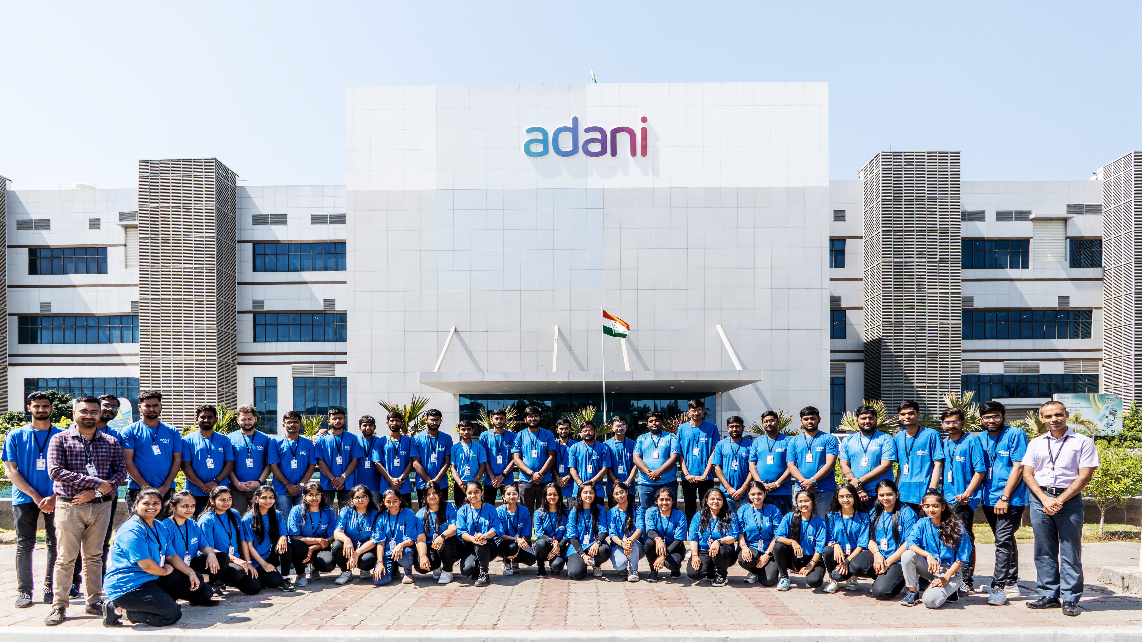 A large group of Adani employees in blue shirts poses for a photo in front of the Adani building.