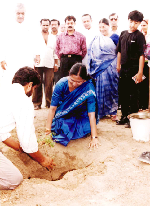 A man and a woman in traditional attire plant a sapling, surrounded by a group of observers.
