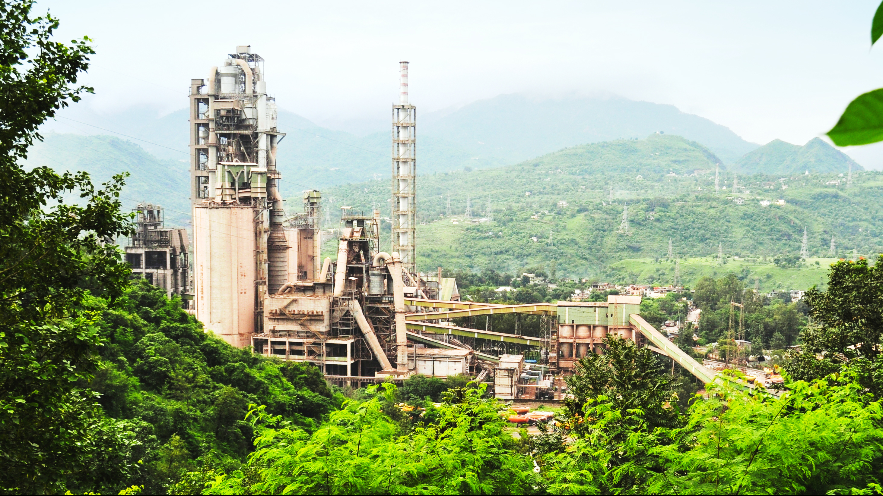 Industrial cement plant complex nestled amidst lush green mountains with a hazy sky.