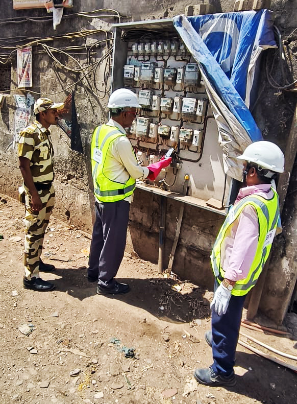Three workers, two in safety vests and helmets, inspect a large outdoor electrical meter panel on a dusty street.