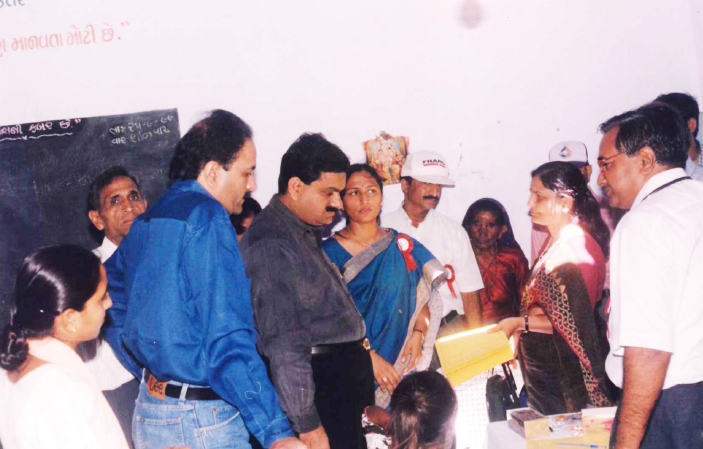 A group of people, including men and women, gathered indoors. Some are looking at a document or object.
