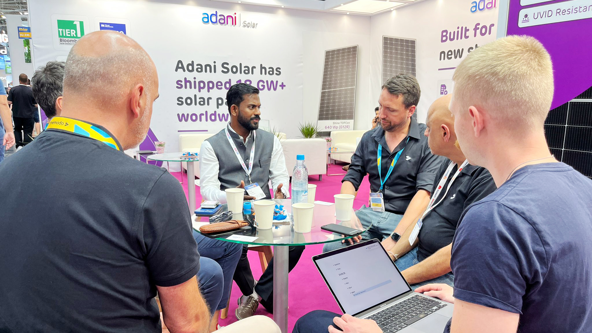 Men discuss solar technology around a table at an Adani Solar booth, with one man using a laptop.