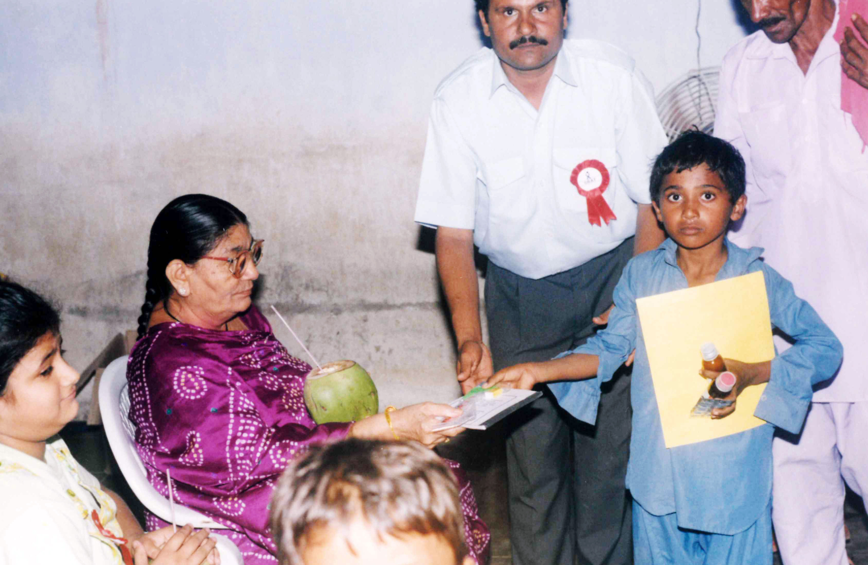 An elderly woman gives an award or certificate to a young boy, assisted by a man, during a community event.