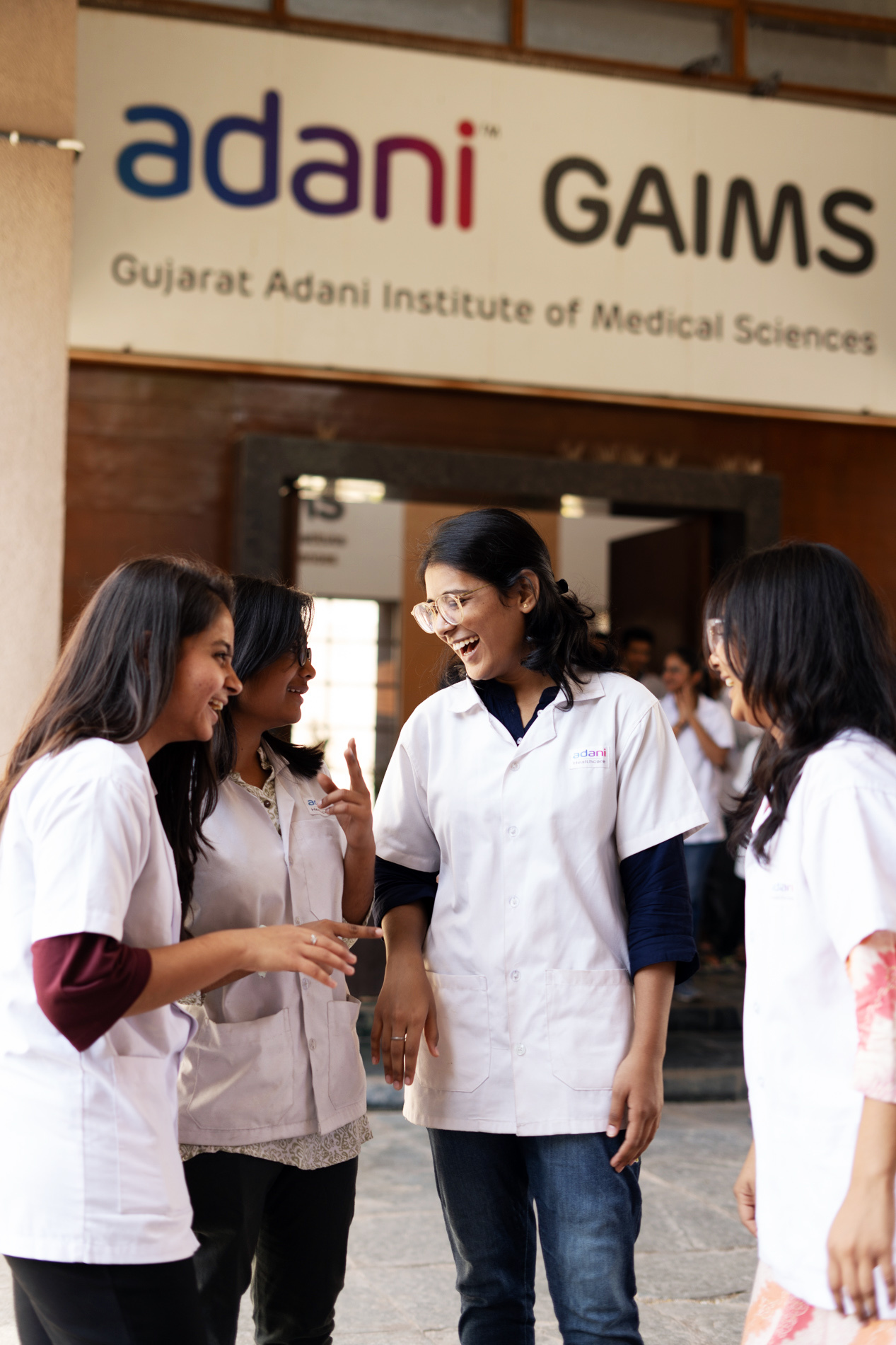 Four female Adani GAIMS medical students in lab coats laughing and talking outside.