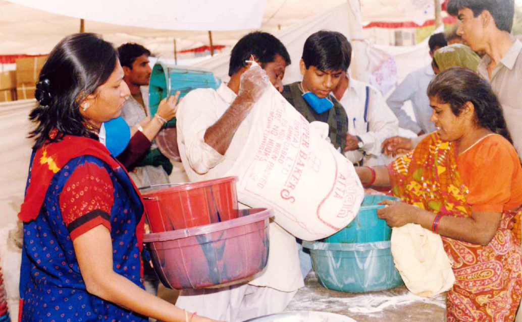 People are distributing flour from a large sack into smaller containers, possibly at a relief camp.