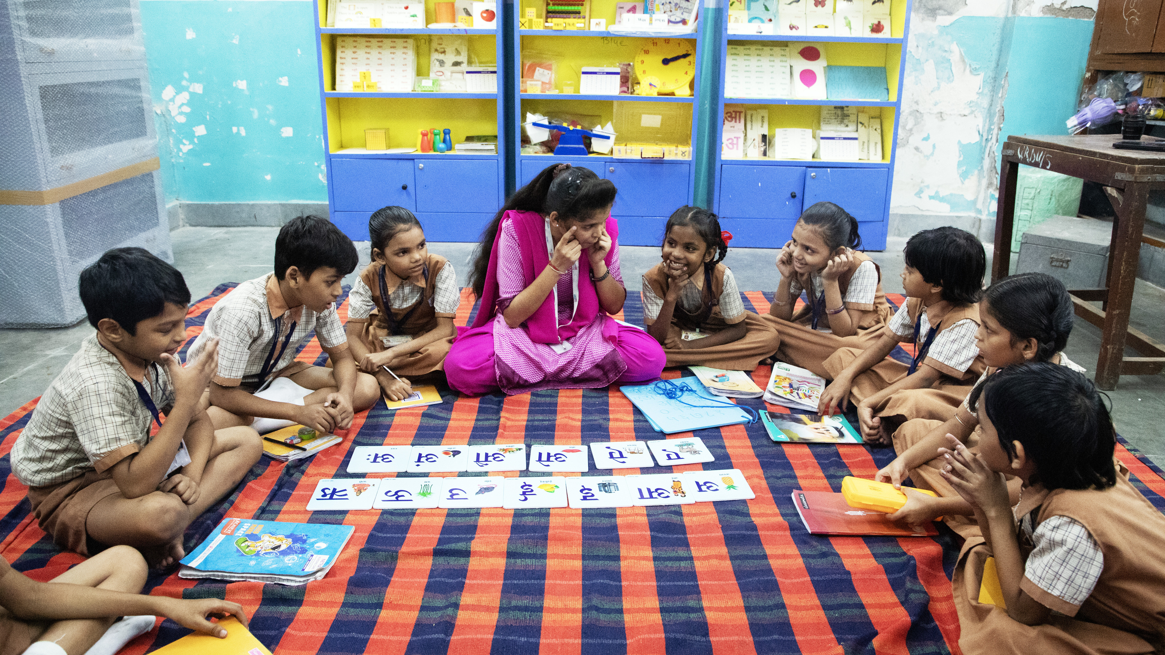 A teacher and students in brown and plaid uniforms sit on a striped mat, learning Hindi letters.