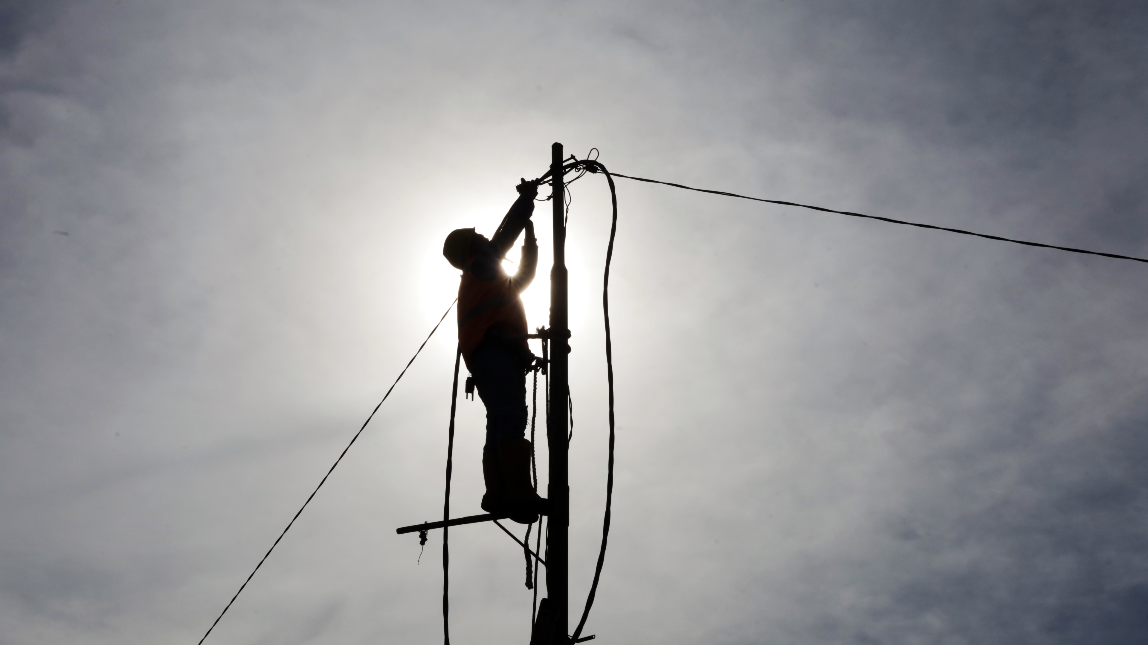 Silhouette of a utility worker on a pole, backlit by a bright sky, performing maintenance on overhead wires.