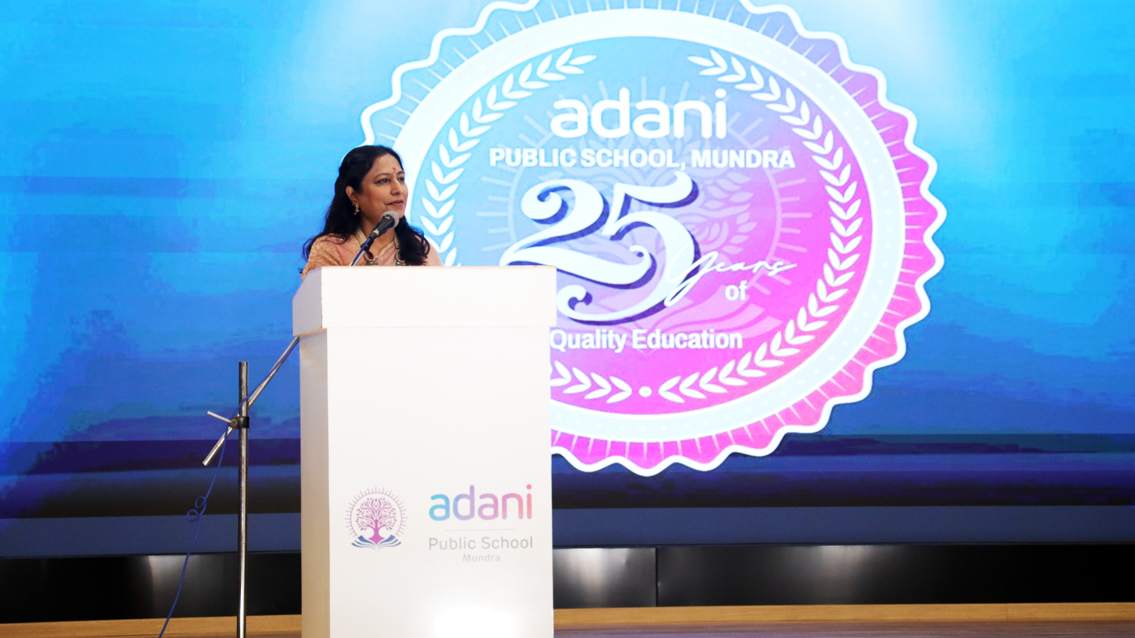 Woman speaking at a podium with Adani Public School 25th anniversary logo on a large screen.
