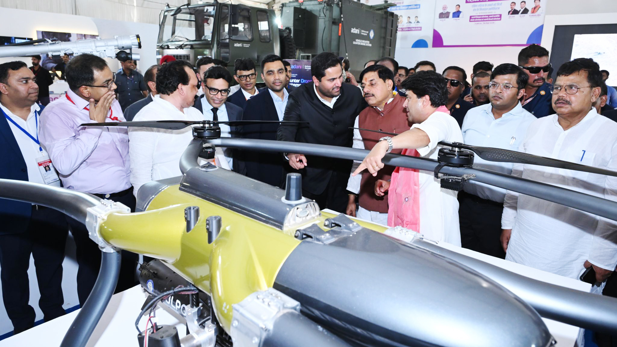 A group of men examining a large drone model at an exhibition. Some wear suits, some traditional attire.