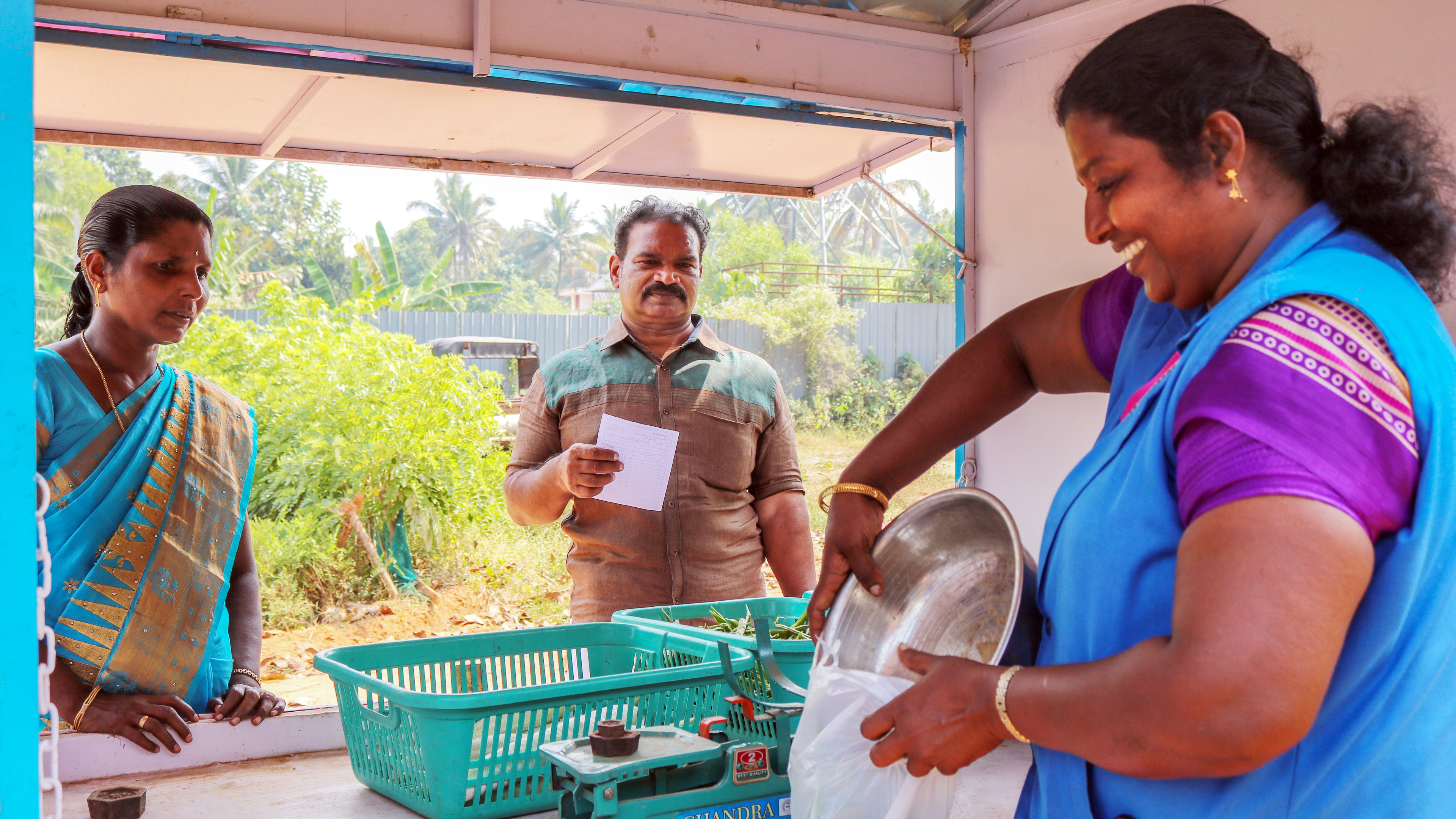 Three people at a market stall, two customers and a vendor weighing produce for a transaction.
