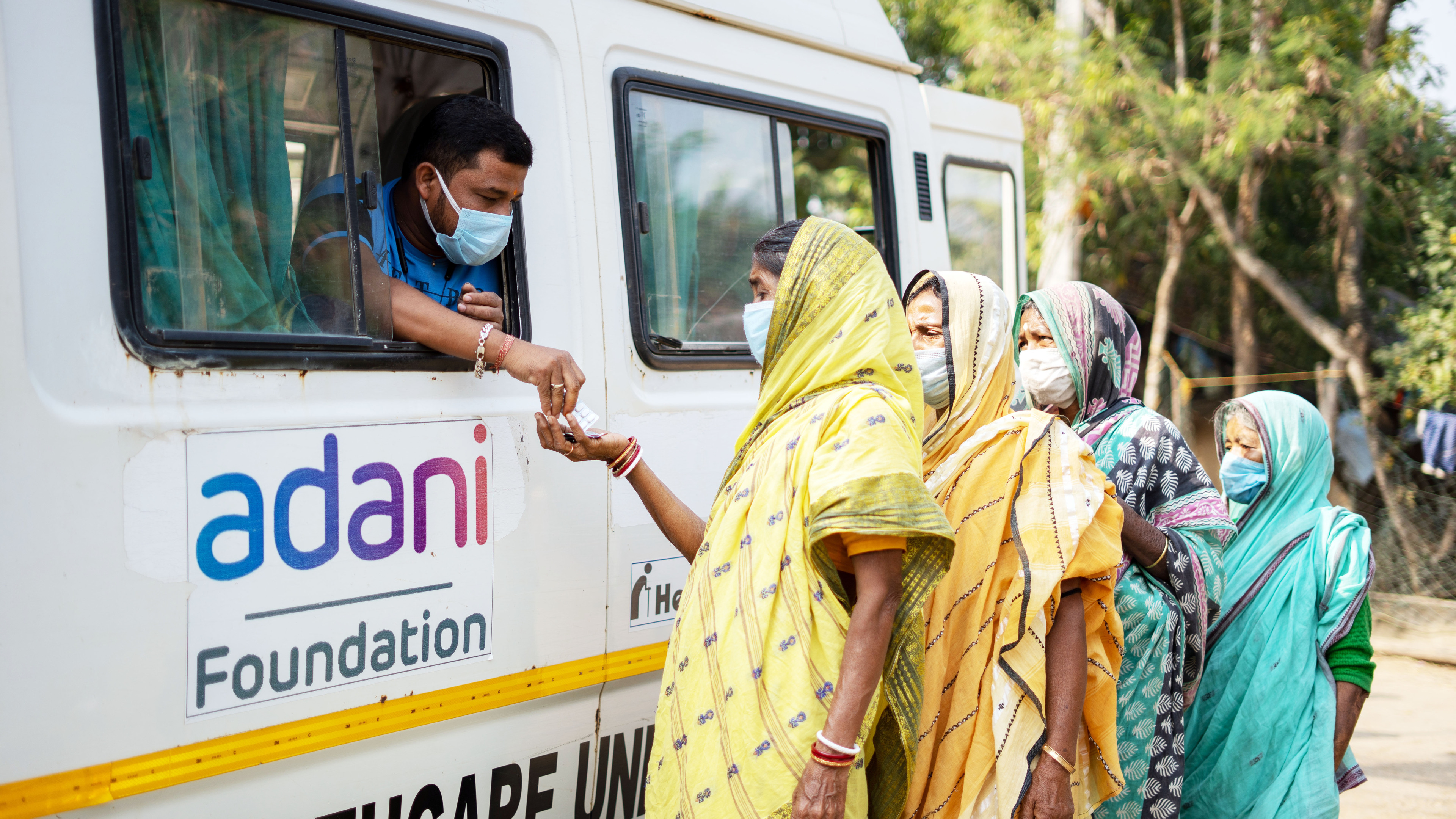 Man in a mask hands medicine from a van to women in masks and saris, representing Adani Foundation.