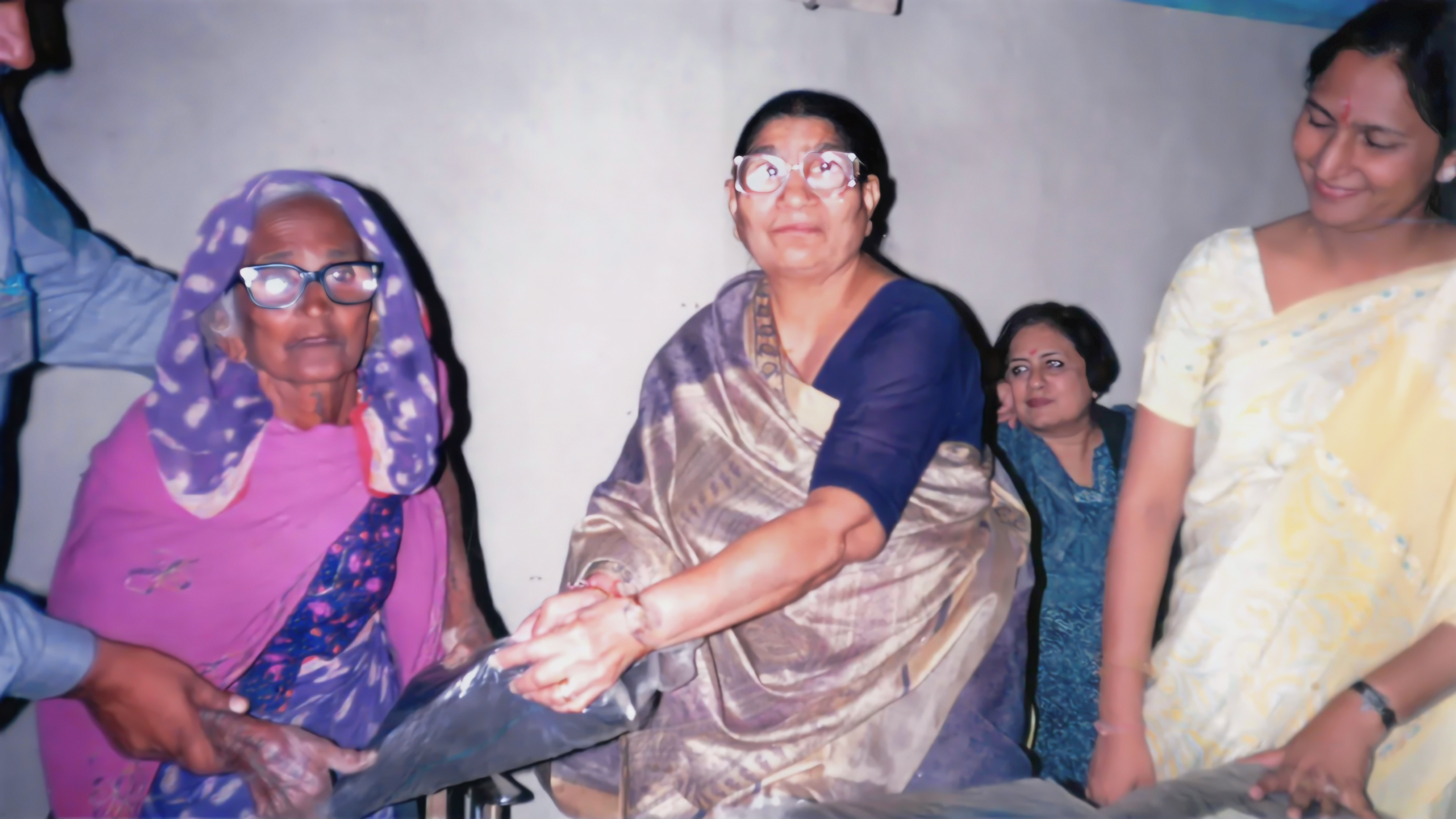 Four women, two wearing glasses, are gathered indoors, with two in the foreground exchanging an item.