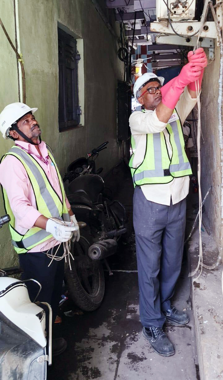 Two electricians in hard hats and safety vests are working with electrical wires in a narrow alley.