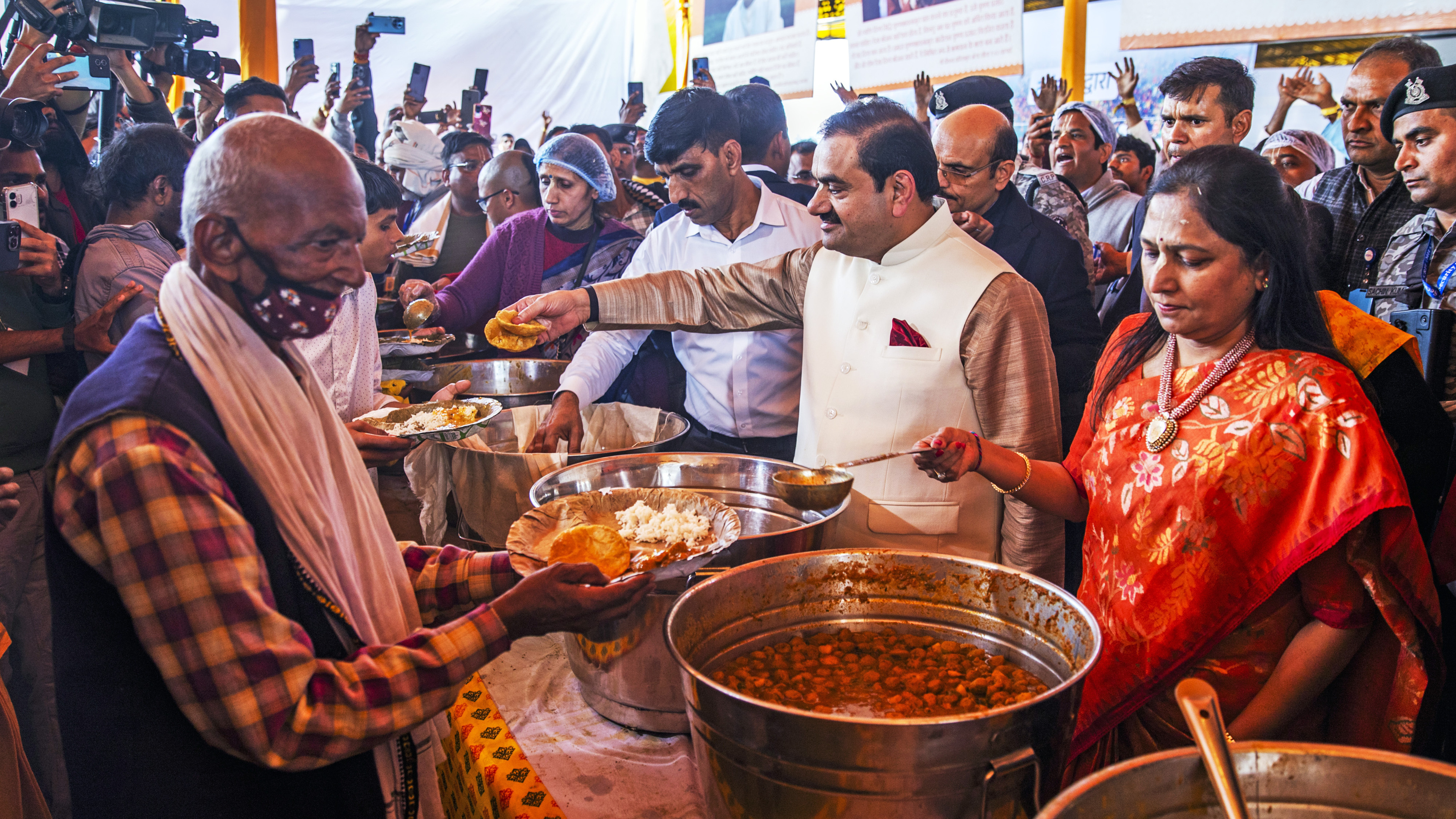 Gautam Adani and a woman serve food from large pots to an old man, amidst a crowd.