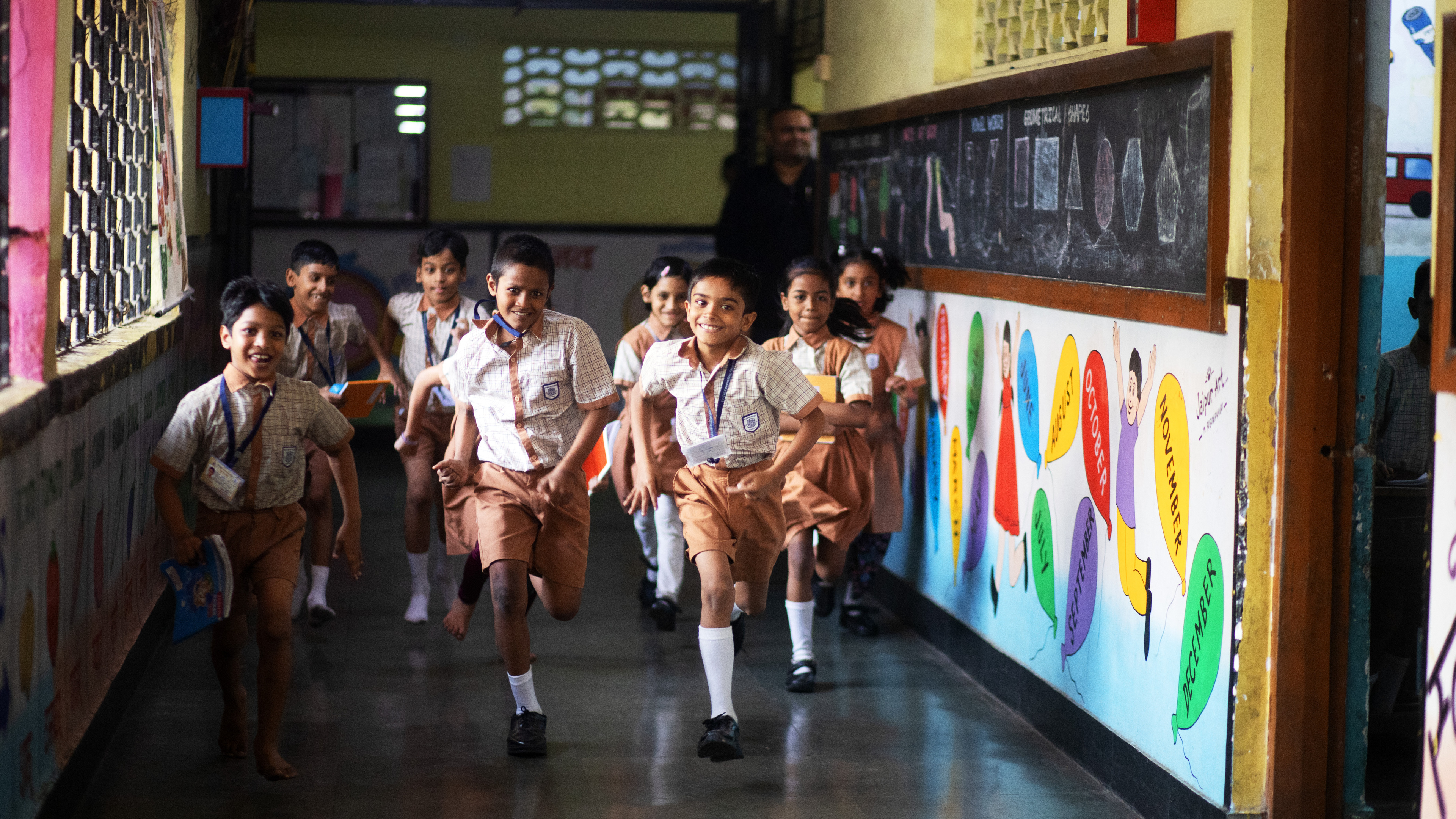 Schoolchildren in uniform run down a brightly lit hallway, smiling and playing with books in hand.