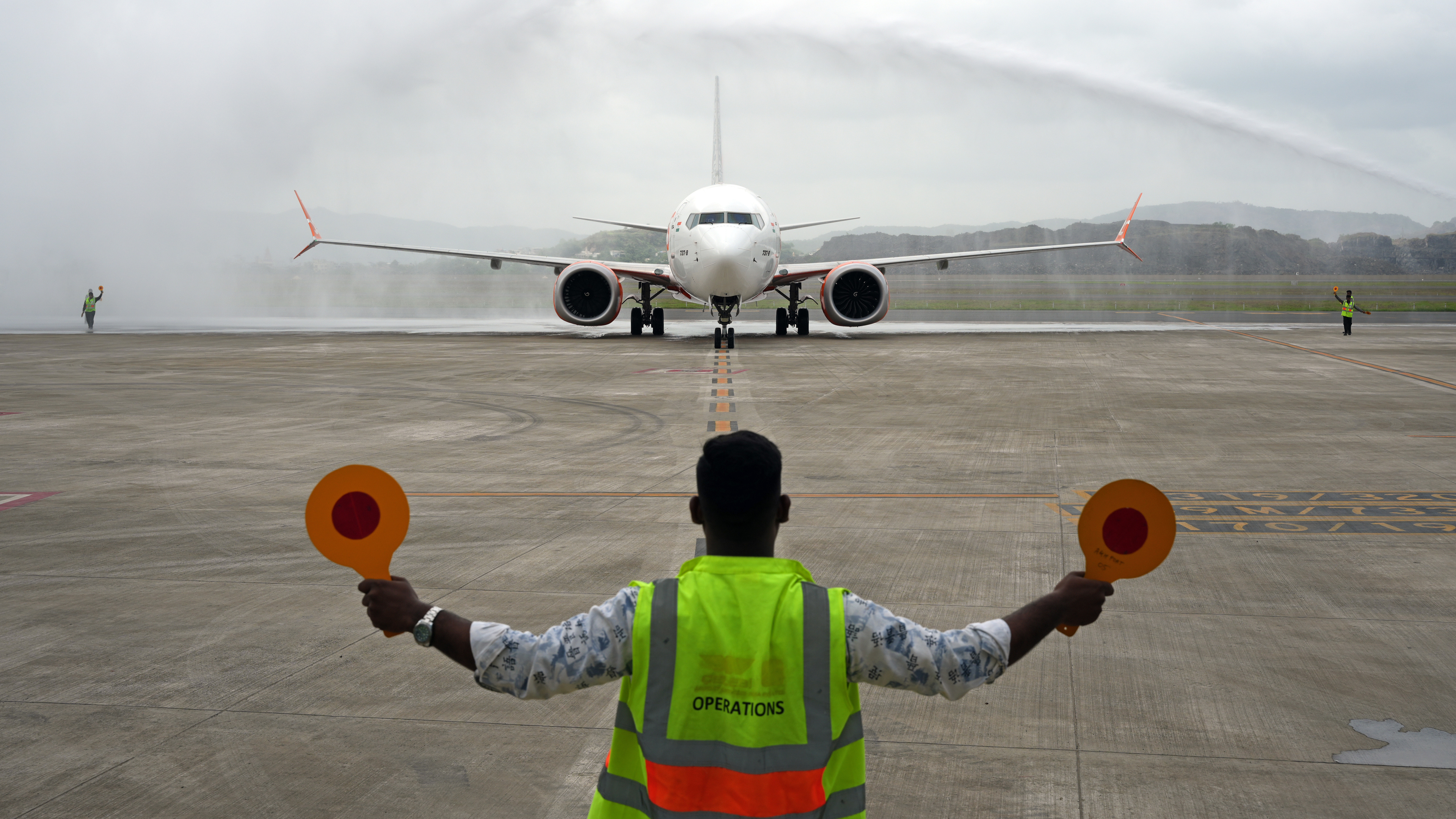 Ground staff directs an airplane receiving a water cannon salute on a cloudy runway.