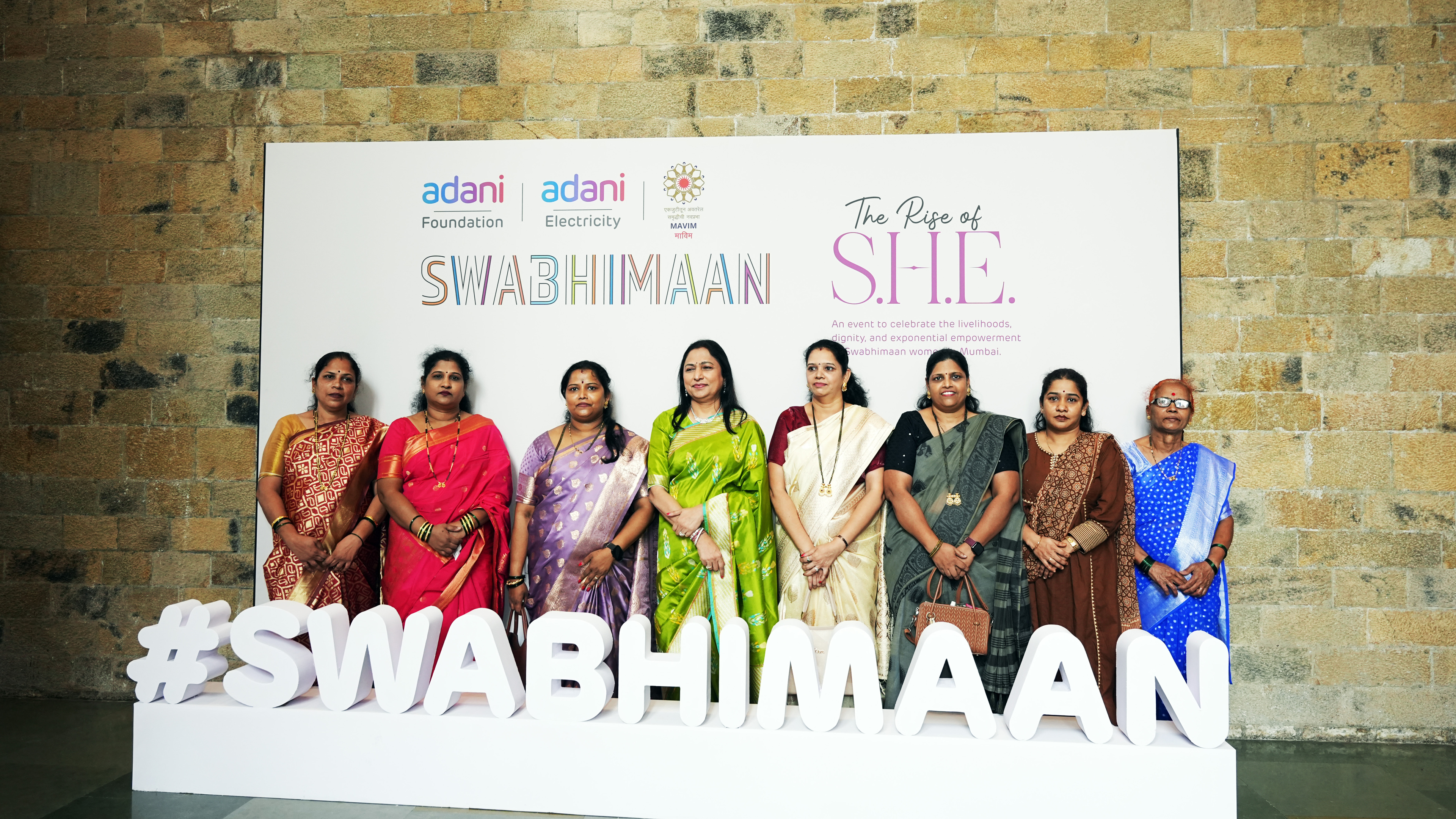 Eight women in traditional Indian attire stand in front of a white banner for the 'SWABHIMAAN' event.