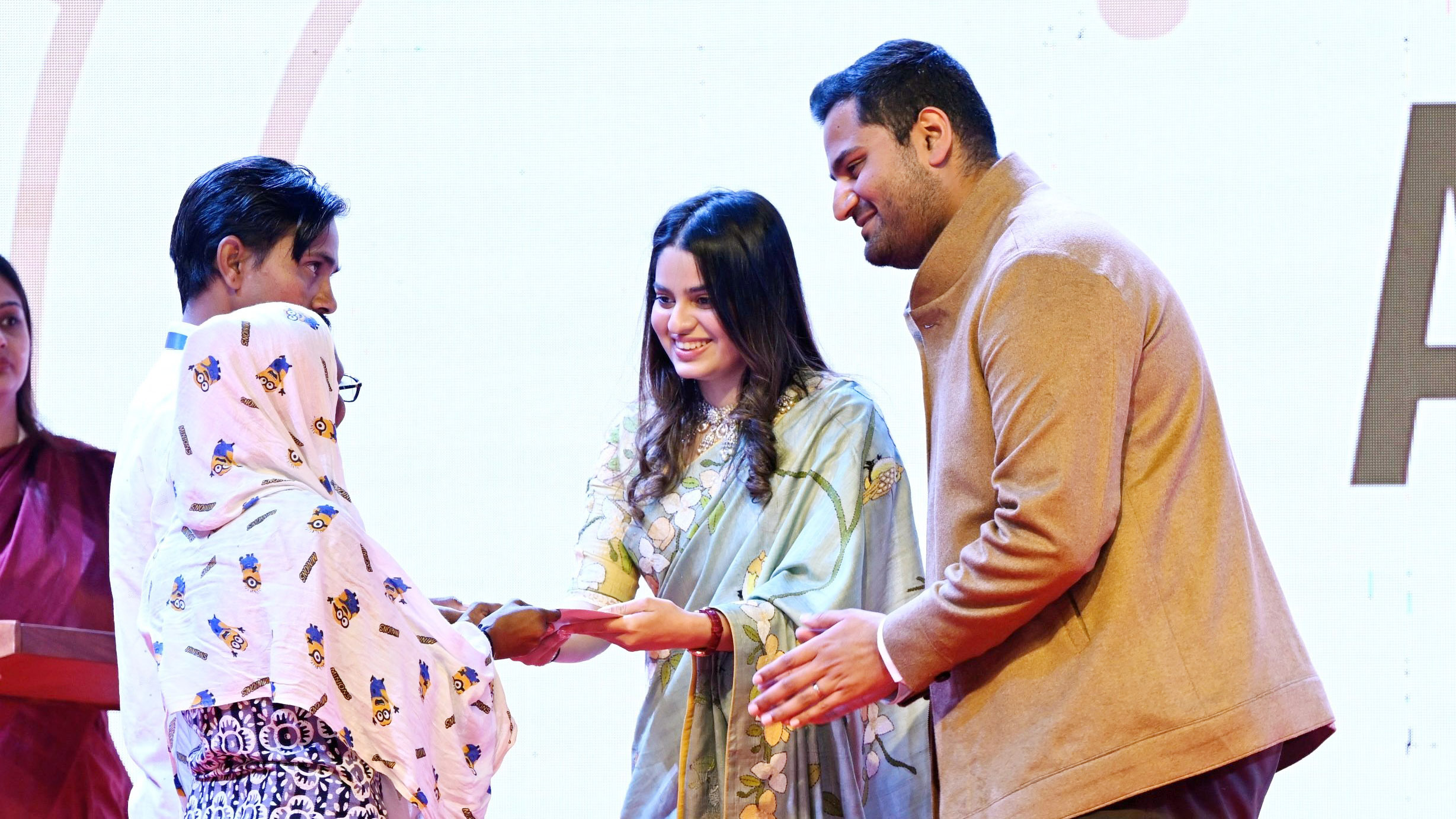 Three people, including a woman in a floral saree, exchange an item at an event.