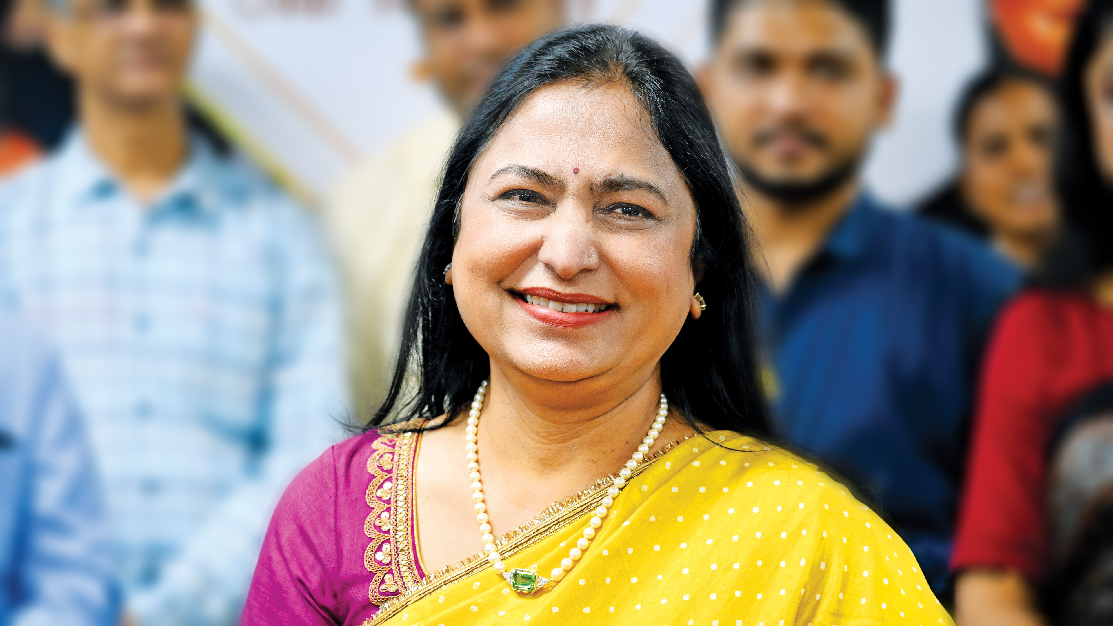 A smiling South Asian woman in a yellow sari and pearl necklace, with blurred people in the background.