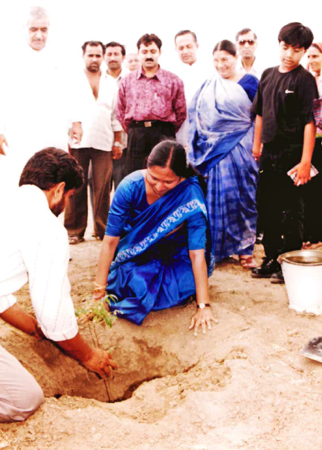 A man and a woman in a blue saree plant a sapling, surrounded by a group of people.