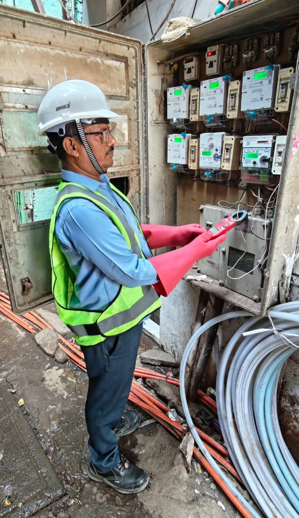 An electrician in safety gear uses a clamp meter to check electrical meters in an open utility box.