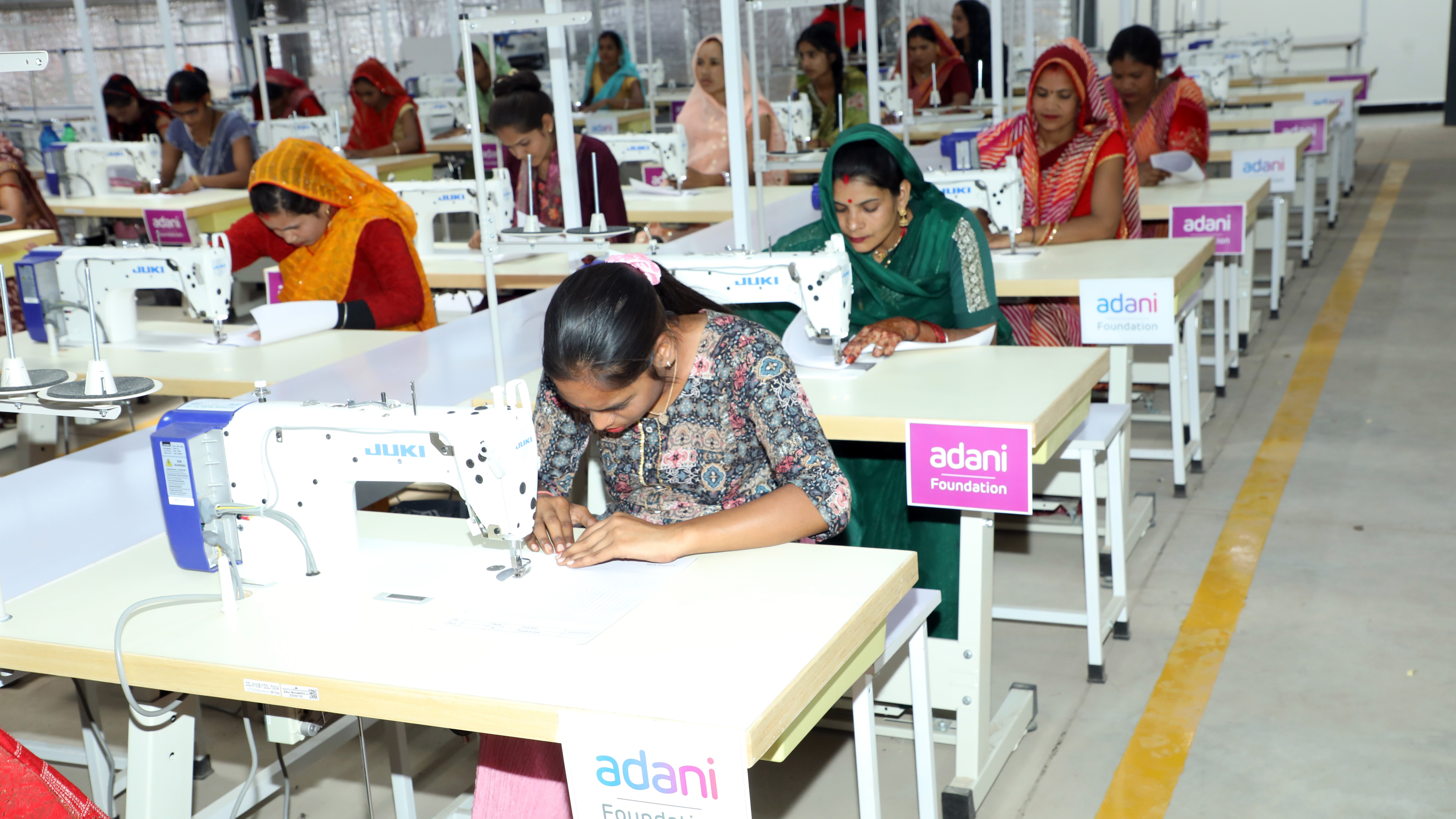 Women in traditional attire operating sewing machines in a garment factory, with "Adani Foundation" logos visible.