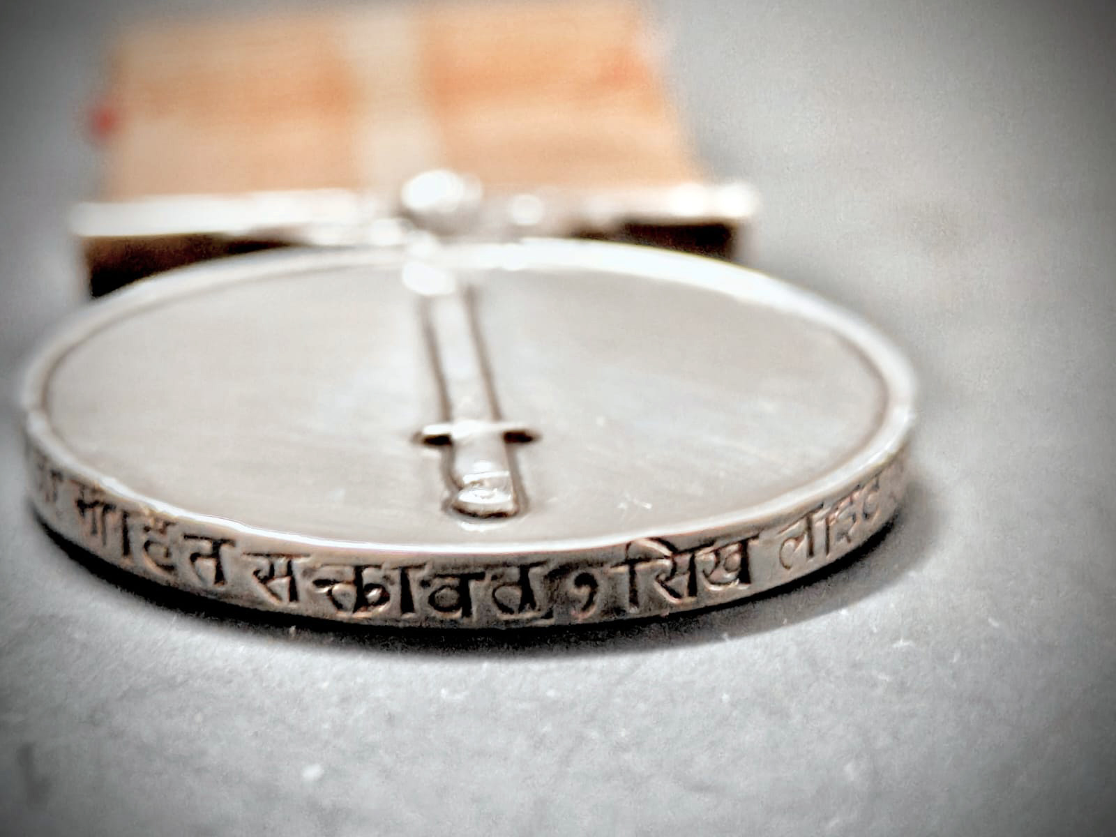 Close-up of a silver medal with Devanagari inscription on its edge and a raised central design, on a blurred background.