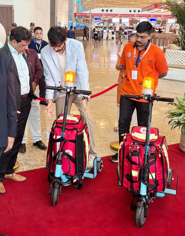 Men examine new medical e-scooters equipped with red bags and flashing lights at an airport terminal.