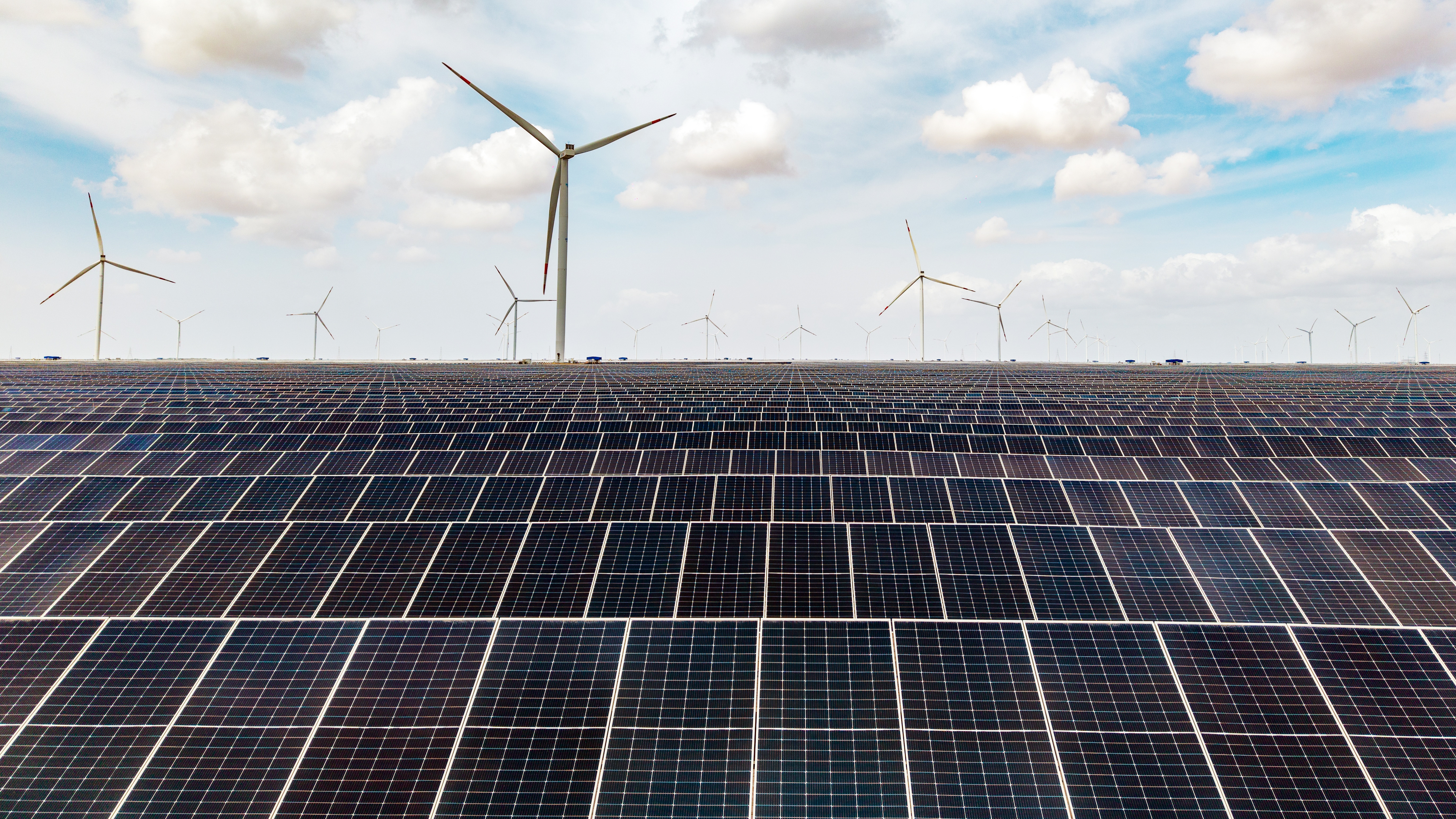 A vast solar farm with rows of photovoltaic panels under a cloudy sky, with numerous wind turbines on the horizon.
