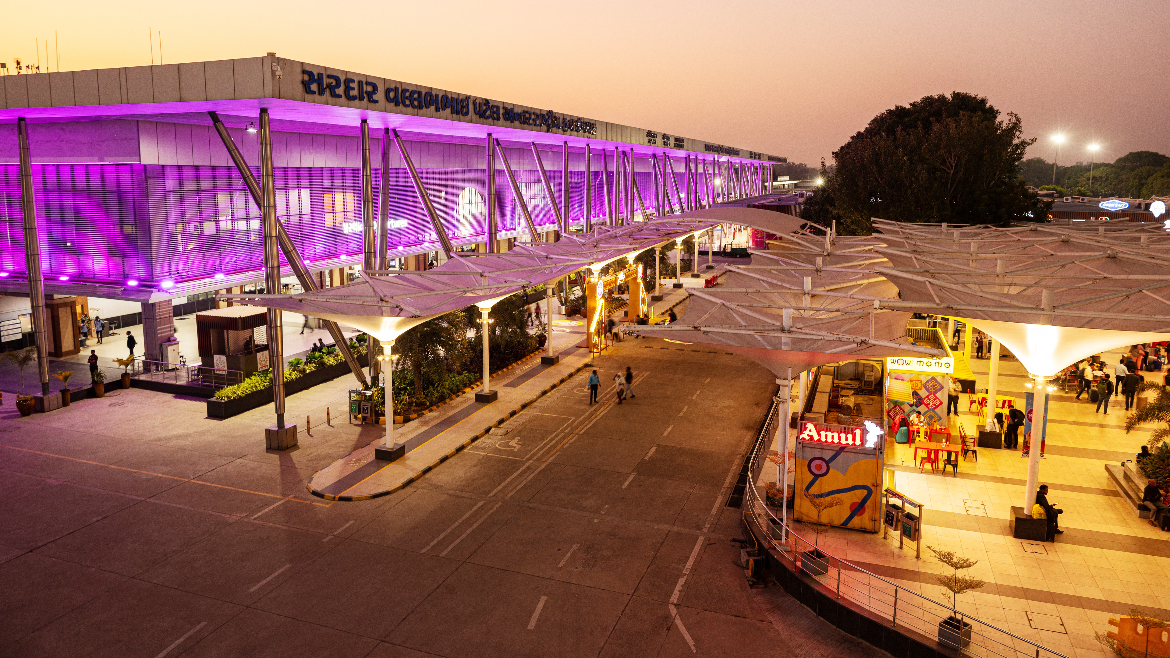 Airport terminal at dusk with vibrant purple lighting, outdoor canopies, shops, and people.