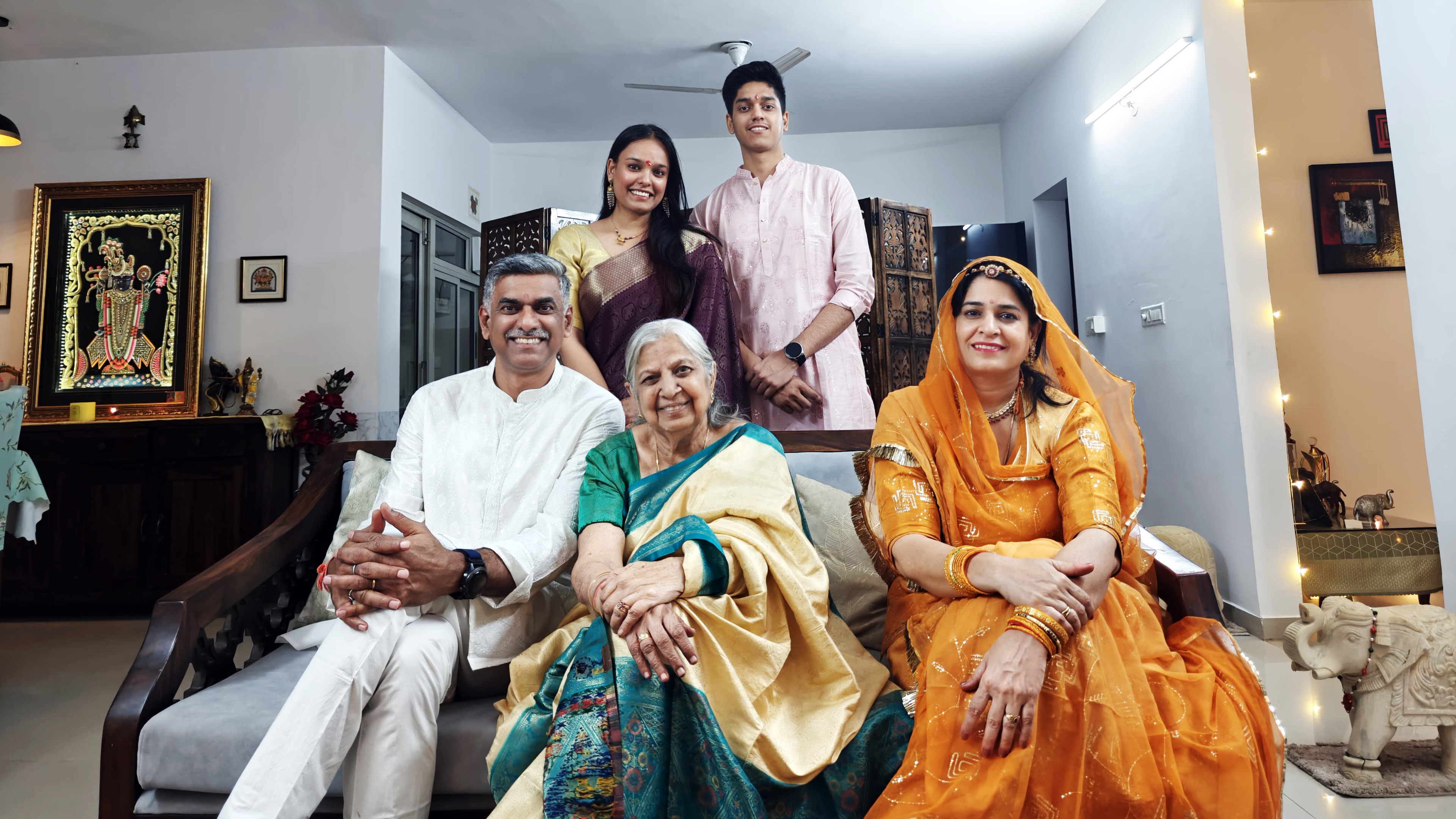 A happy Indian family of five, generations gathered in a living room, smiling in traditional attire.
