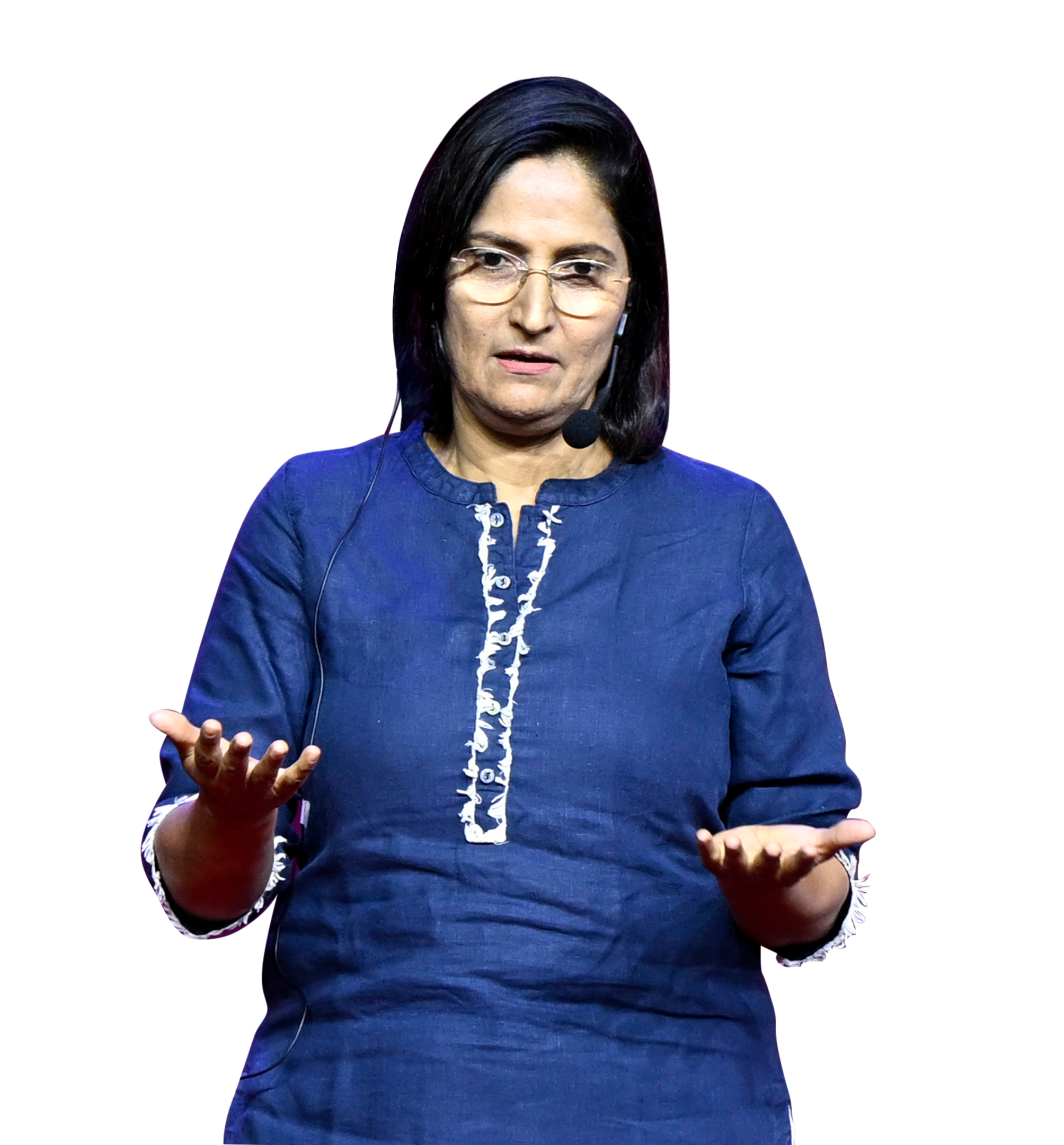 A woman in a blue tunic and glasses speaks into a microphone, gesturing with open hands.