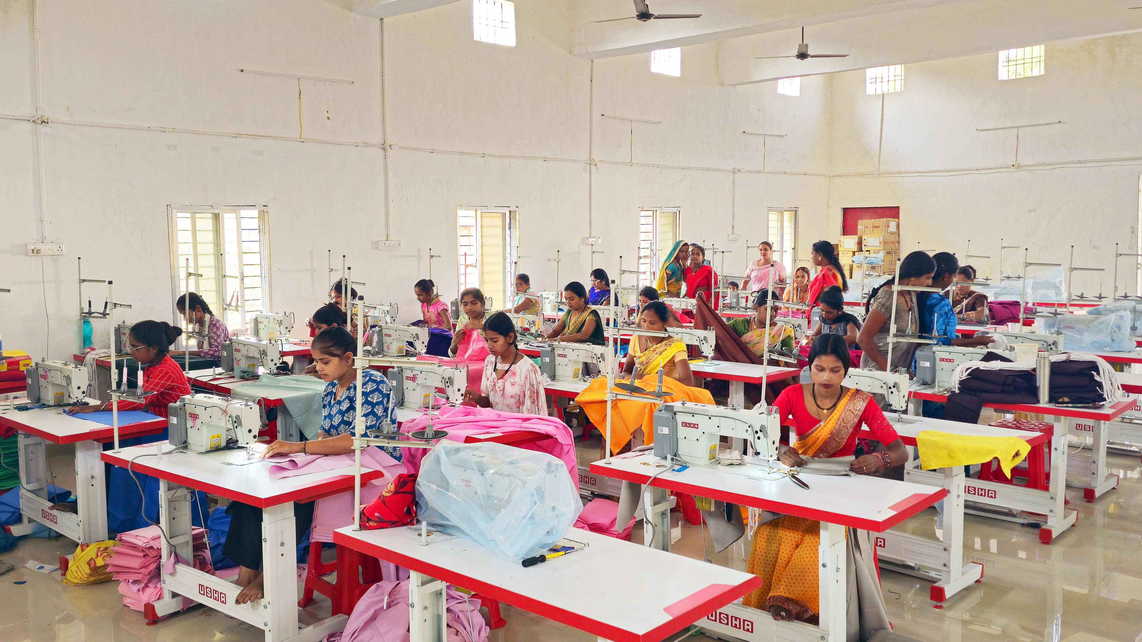 Many women sewing clothes on machines in a bright textile workshop.