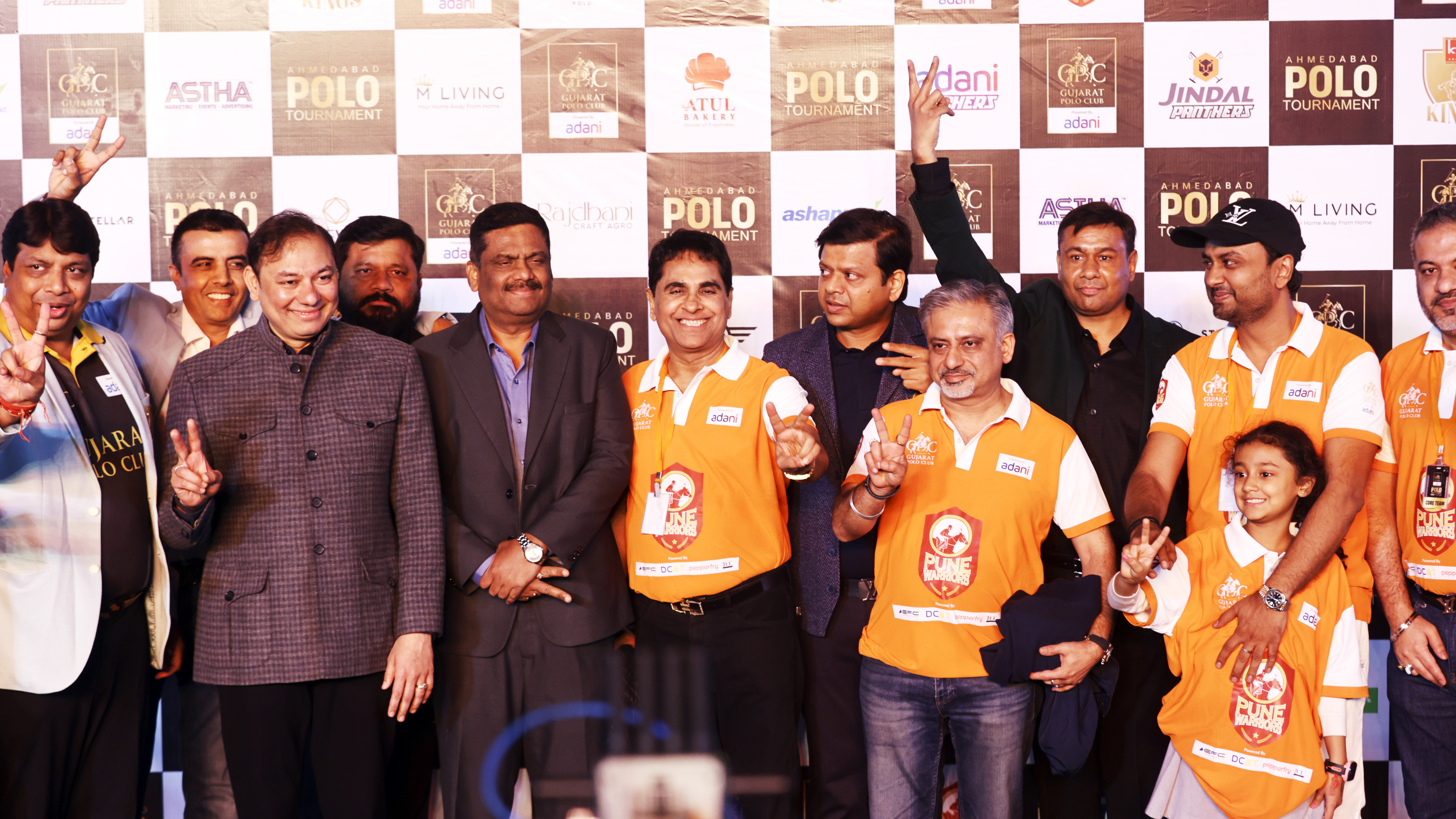A group of men and a girl, many in orange Adani Pune Warriors shirts, pose with V-signs at a polo event.