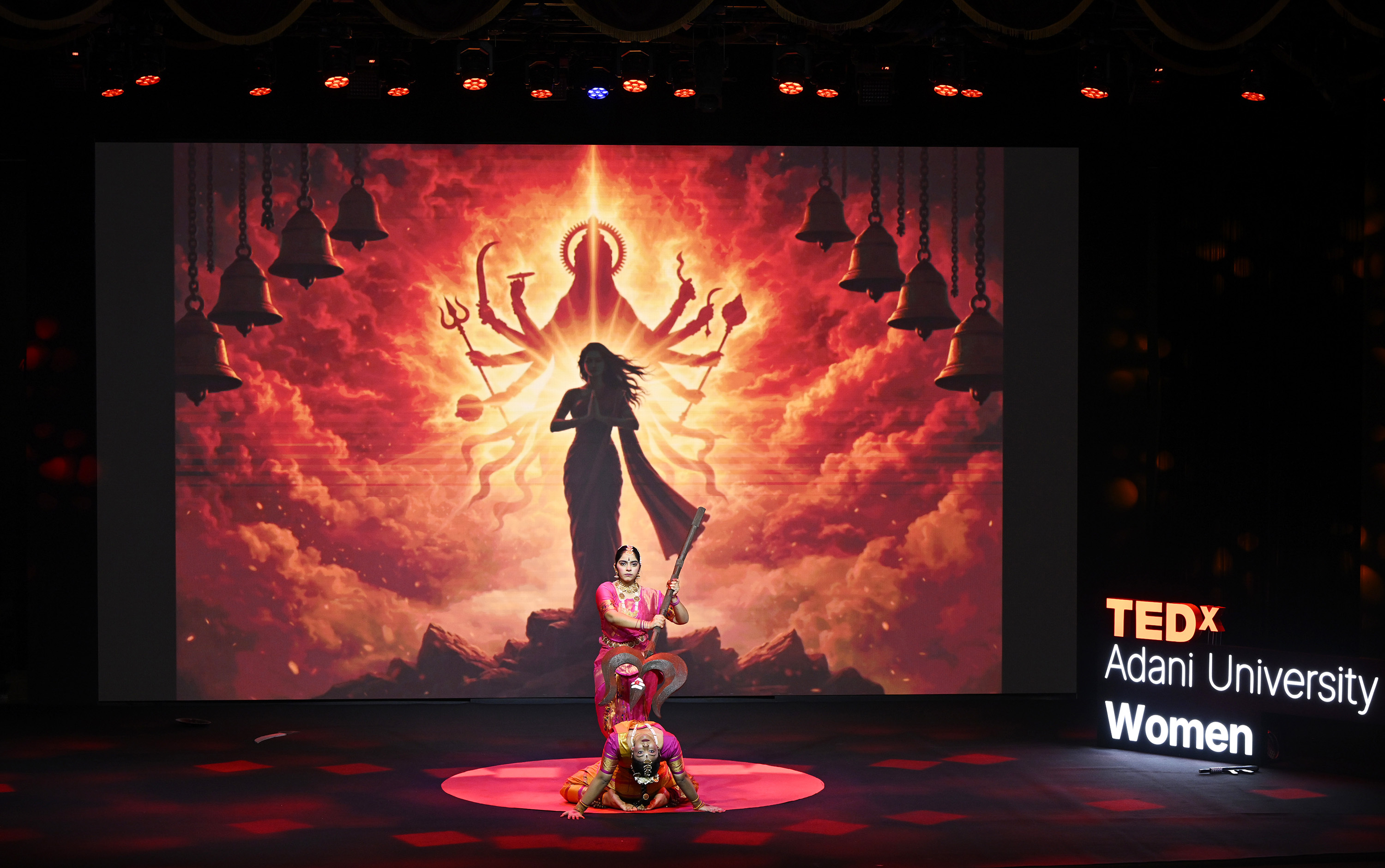 Two dancers perform on stage with a vibrant Hindu goddess projection and "TEDx Women" sign.