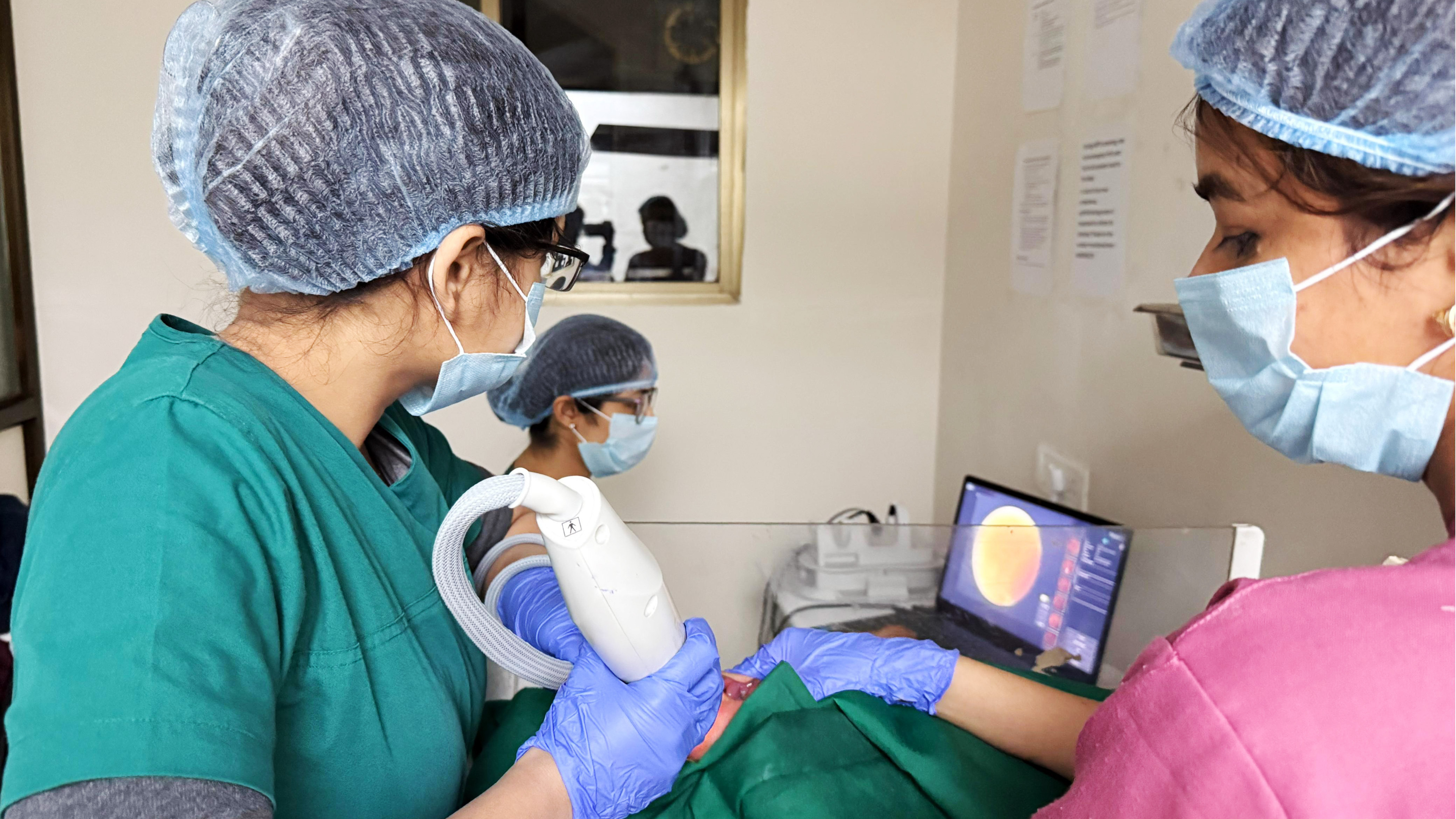 Two medical professionals perform a procedure using a handheld device while monitoring a laptop screen.