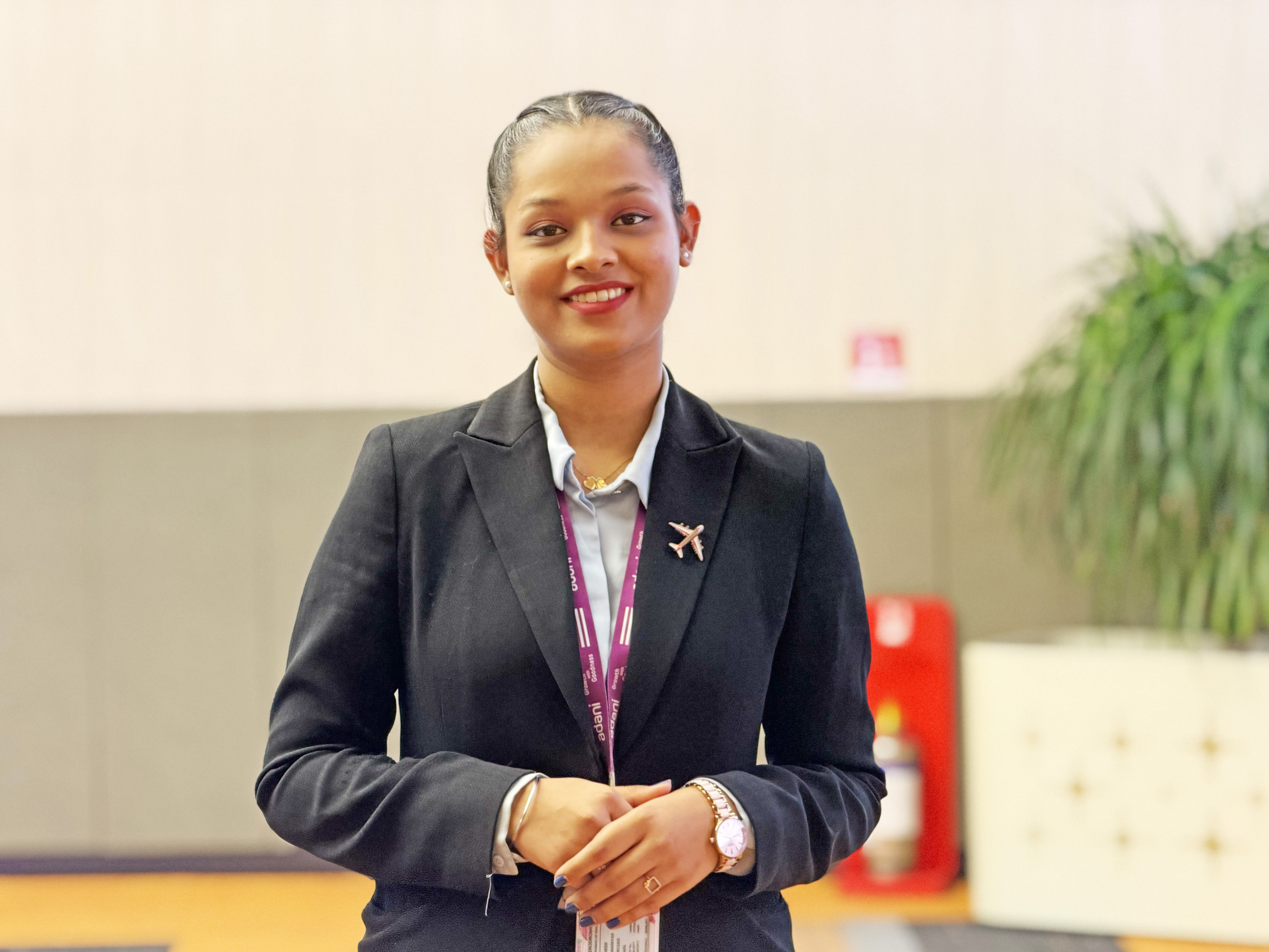 A smiling female airport staff member in a black suit with an airplane pin and ID lanyard.