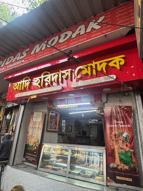 The storefront of Adi Haridas Modak, a sweet shop in Kolkata, with display cases of Indian sweets and a Ganesh Chaturthi banner.