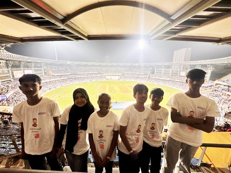 Five young people in Adani t-shirts pose at a brightly lit, crowded cricket stadium.