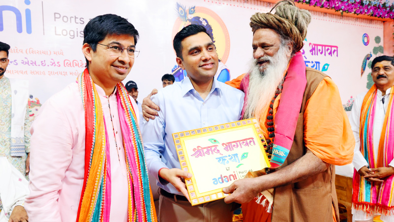 Two men present an 'Adani Srimad Bhagavat Katha' frame to a sadhu at an event.