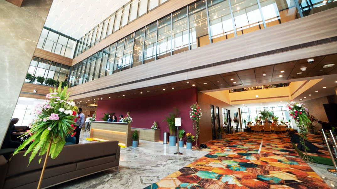 Spacious modern lobby with a reception desk, colorful carpet, high glass walls, and flower arrangements.