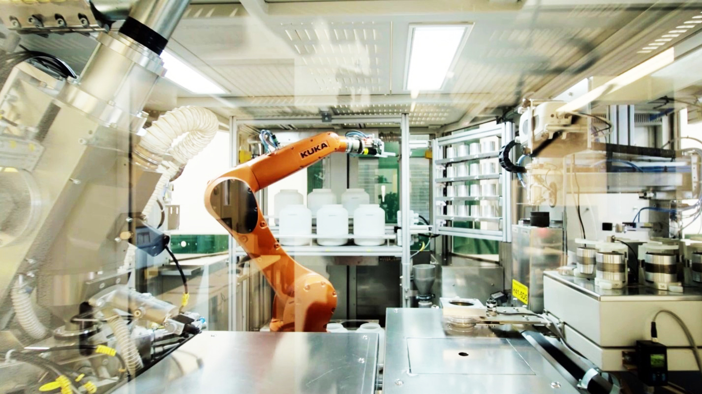 Orange KUKA robotic arm in a sterile, automated industrial or laboratory setup with containers.