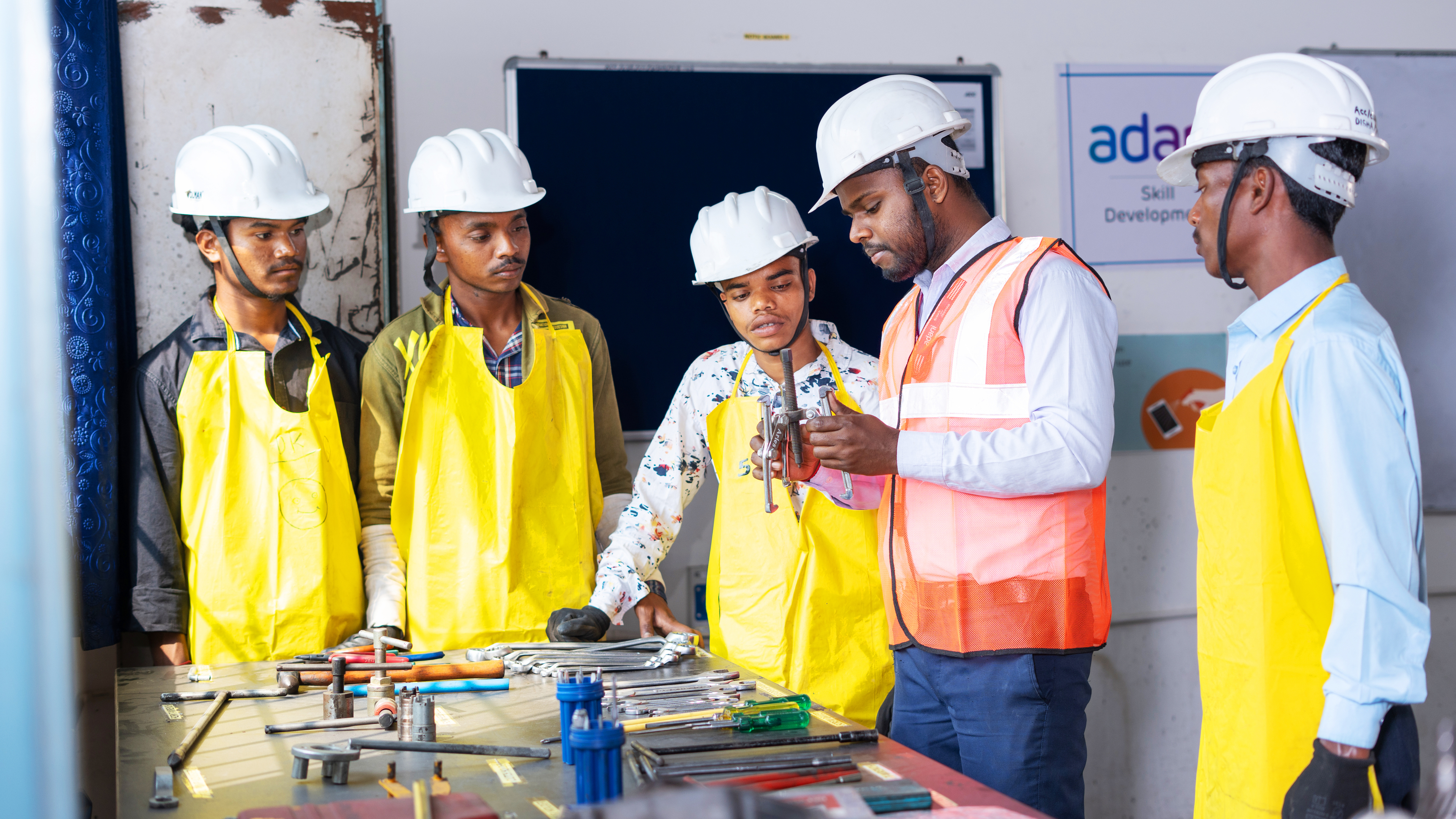 Five men in hard hats and aprons learning about tools in a workshop.