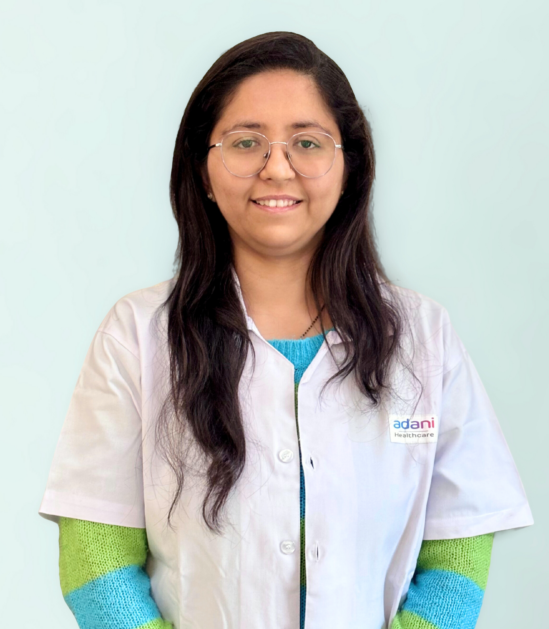 Smiling woman wearing glasses, white lab coat with "Adani Healthcare" logo, and colorful sweater.