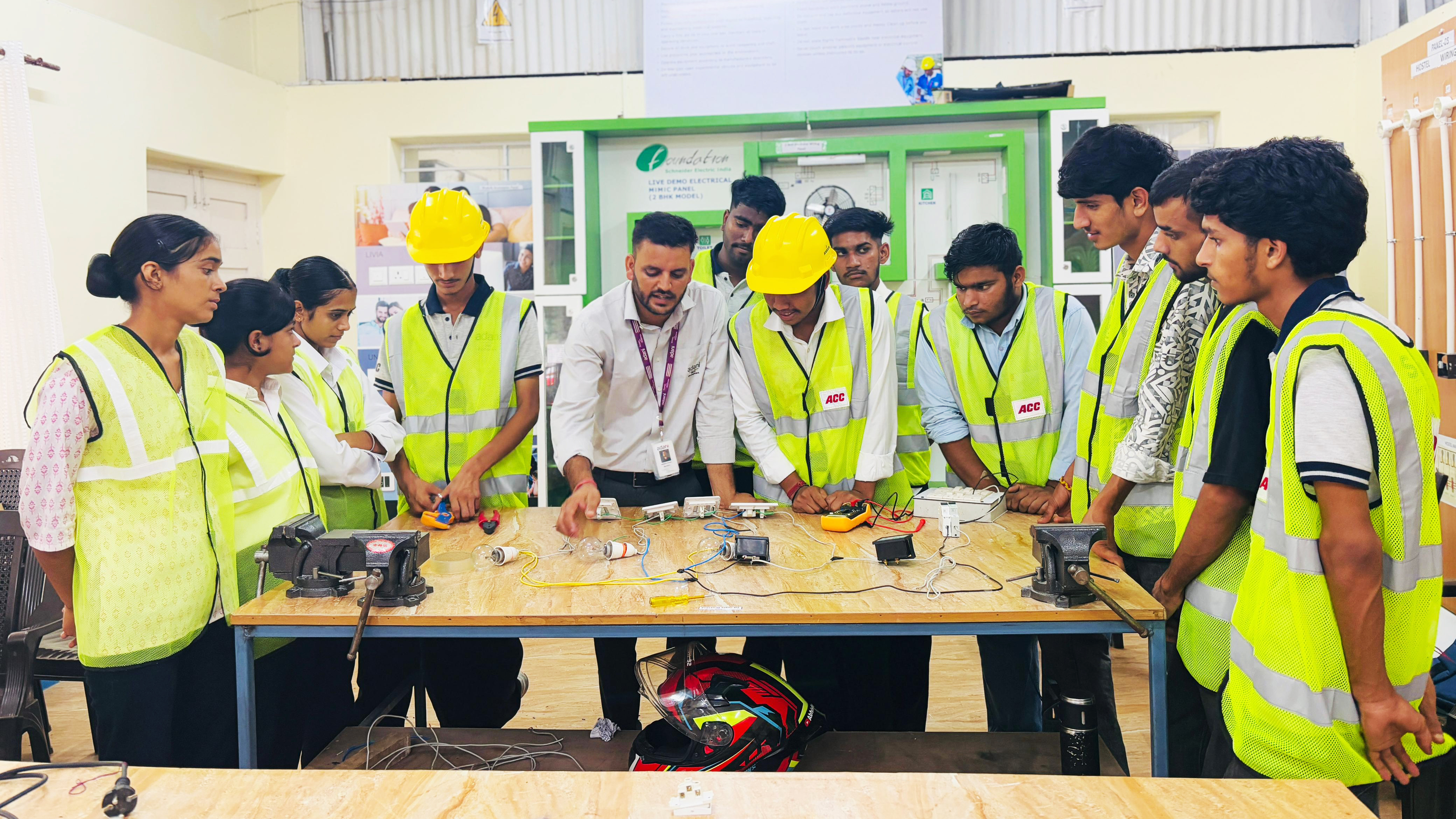 Instructor demonstrates electrical wiring to students wearing safety vests and hard hats.