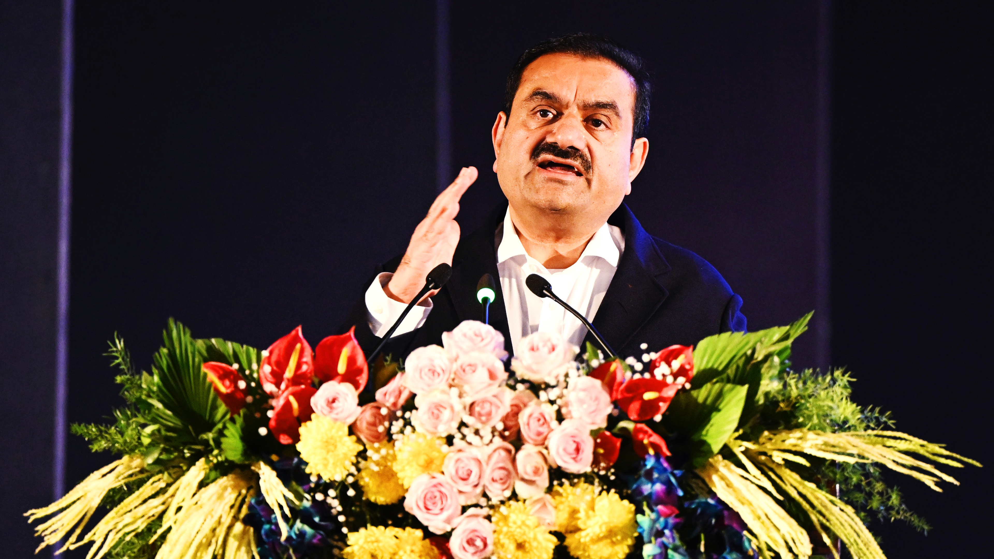 Gautam Adani speaking at a podium adorned with flowers, gesturing.